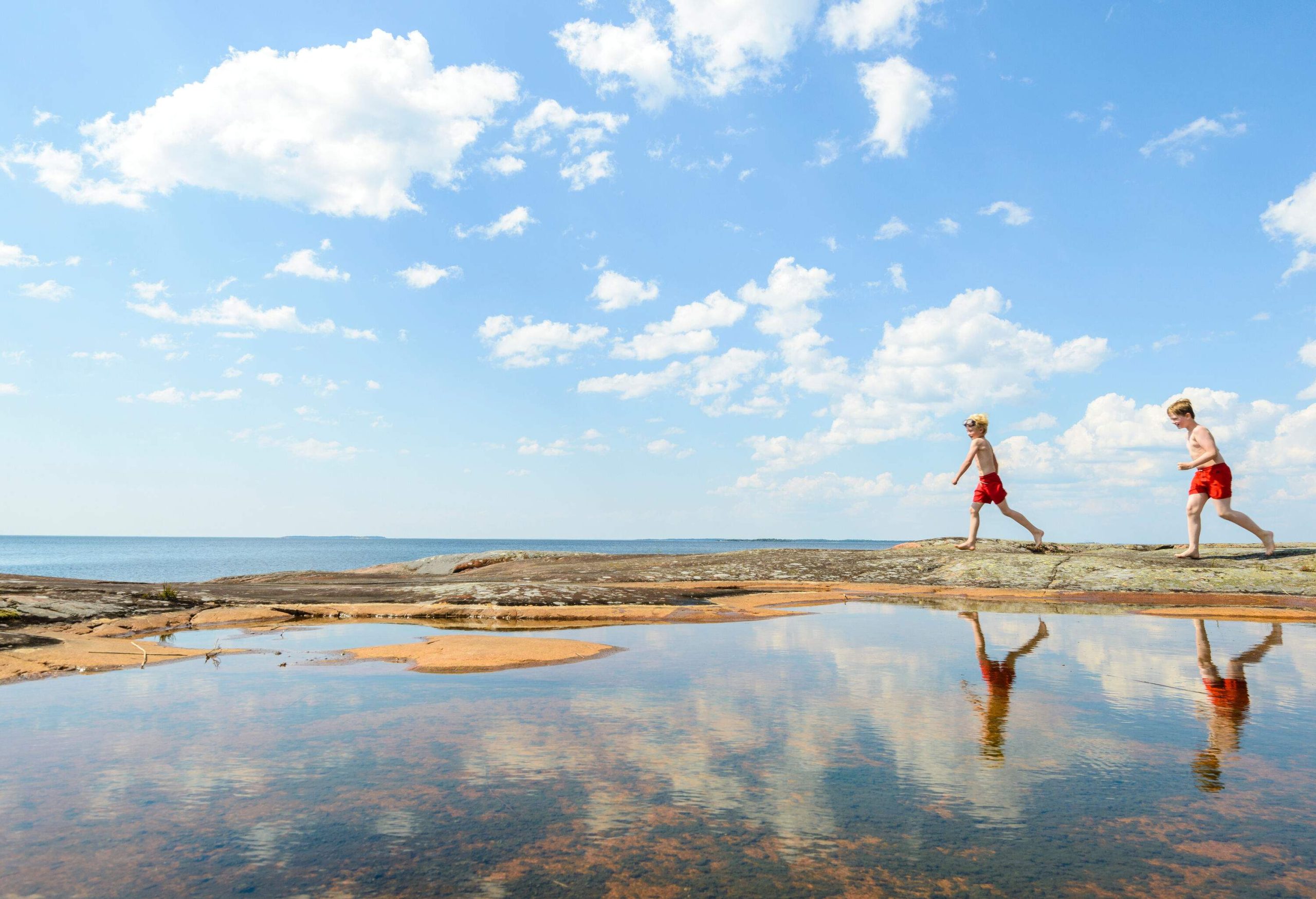 Two kids walk on flat rocks next to a beach's shallow waters.