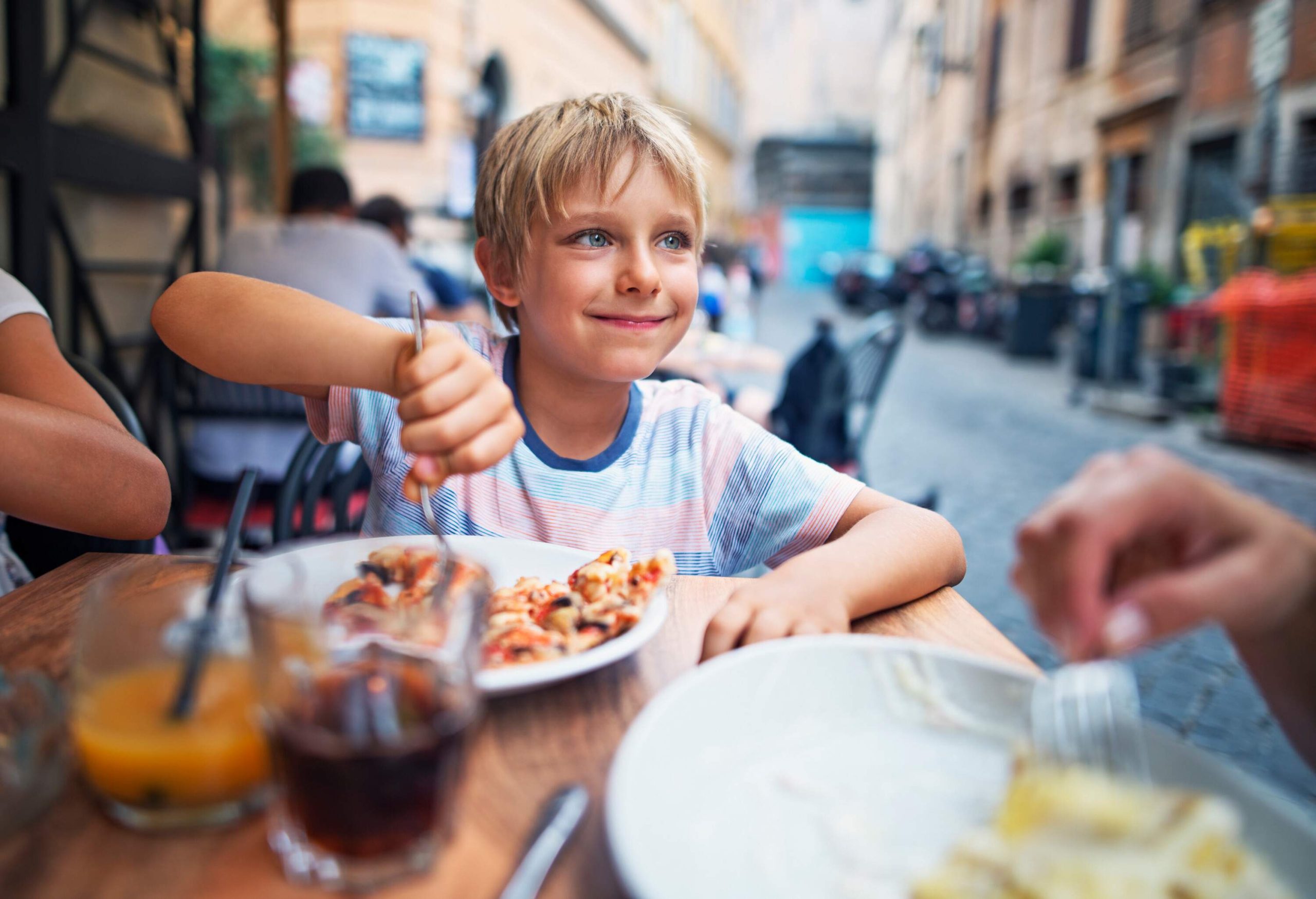 A young boy with blonde hair eating pizza with a fork at an outdoor restaurant.