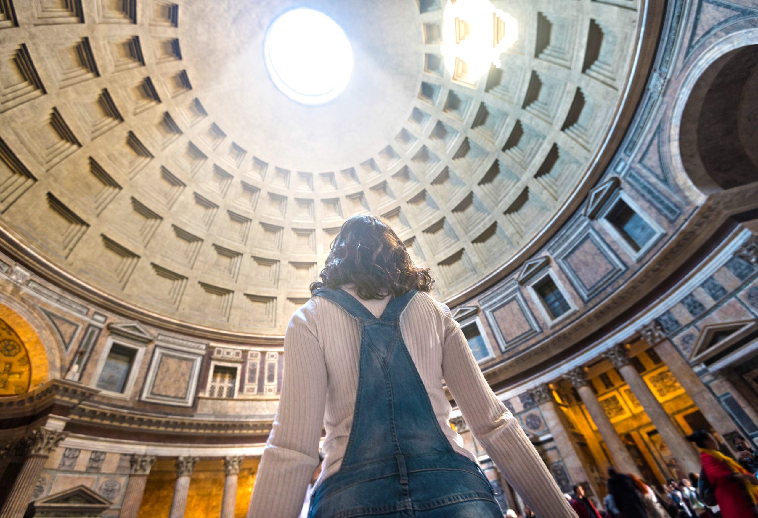 A woman is standing inside a building with a coffered dome with light passing through the central oculus.