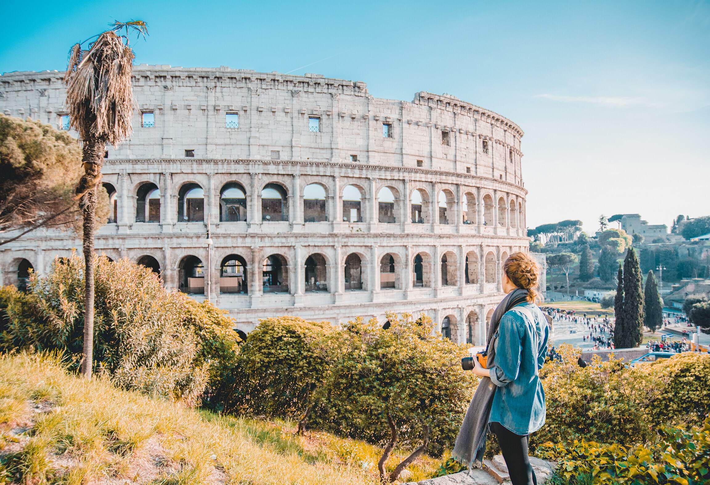 A female tourist with a camera admiring the beauty of the Colosseum from a hill.