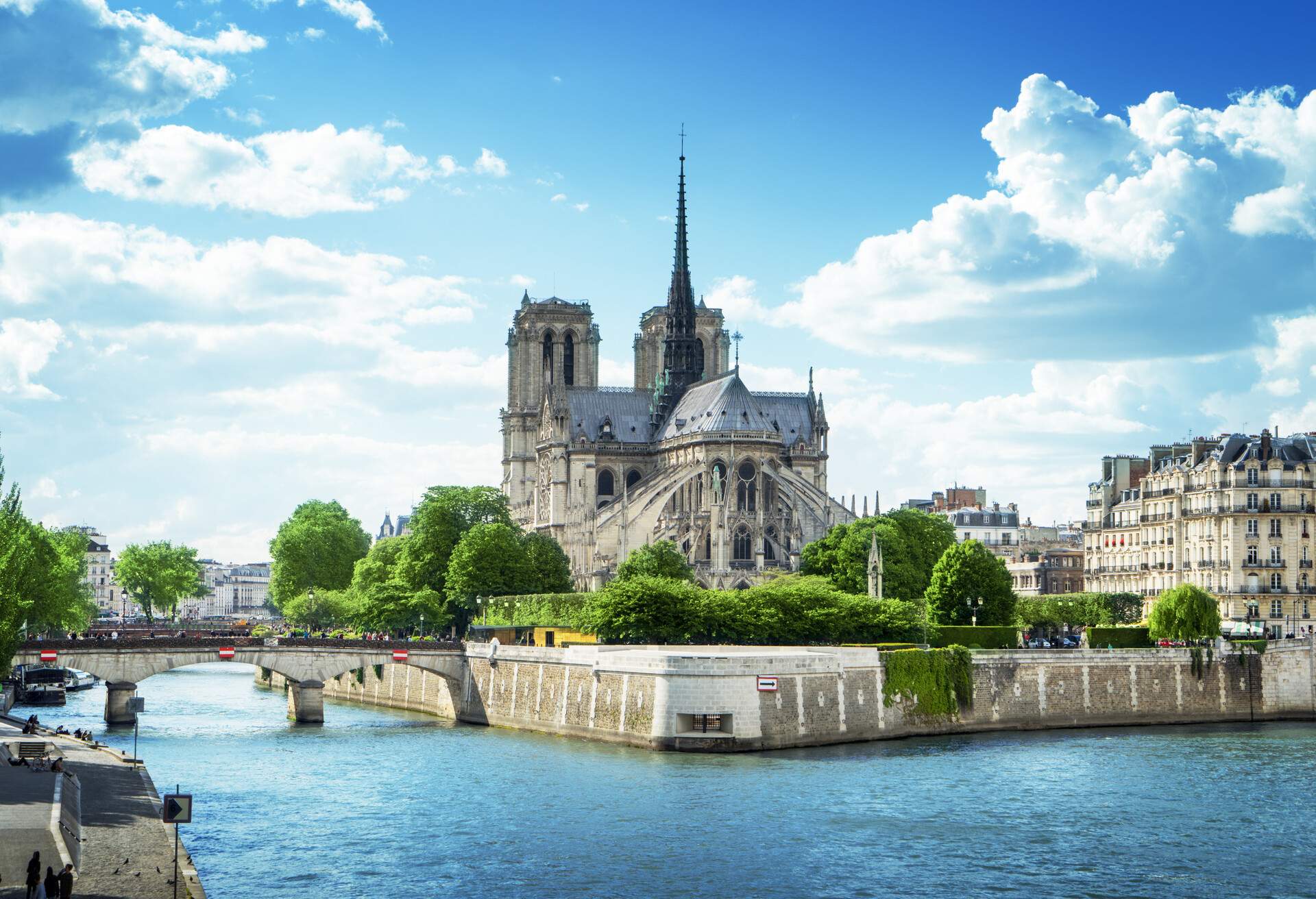 A medieval cathedral with a spectacular ornate spire surrounded by lush trees accessed through a bridge in the middle of a river.