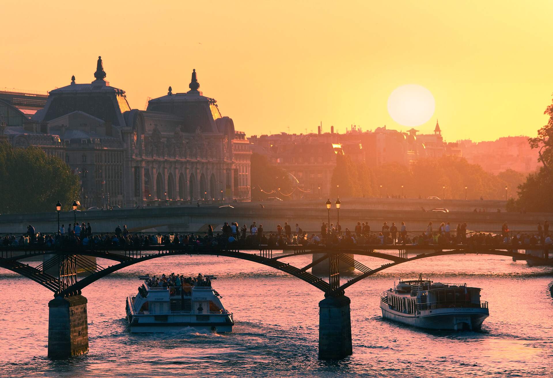 Loaded ferry boats sailing in a river with an arch bridge packed with people at sunset.