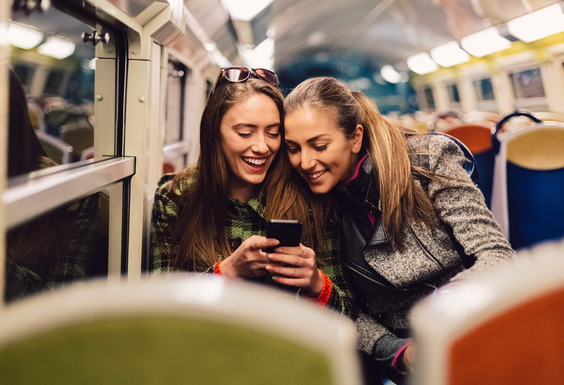 Two happy women sit on a train and look at a mobile phone.