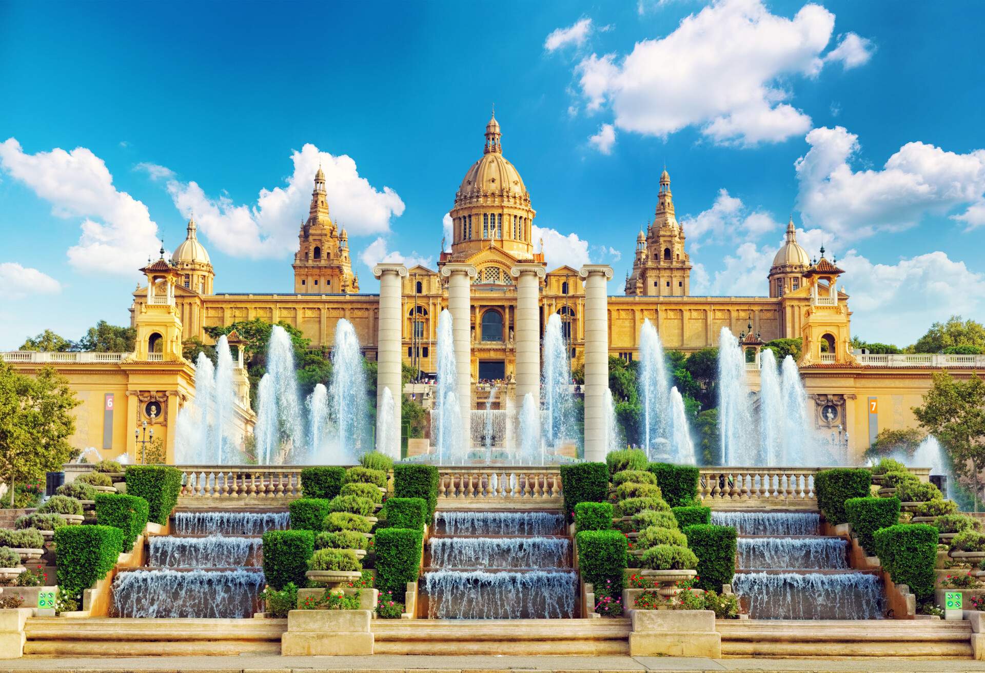 A neoclassical style museum with domed towers and a water fountain. There is also a three-tiered waterfall in front lined with box hedges between the stairs leading to the building.