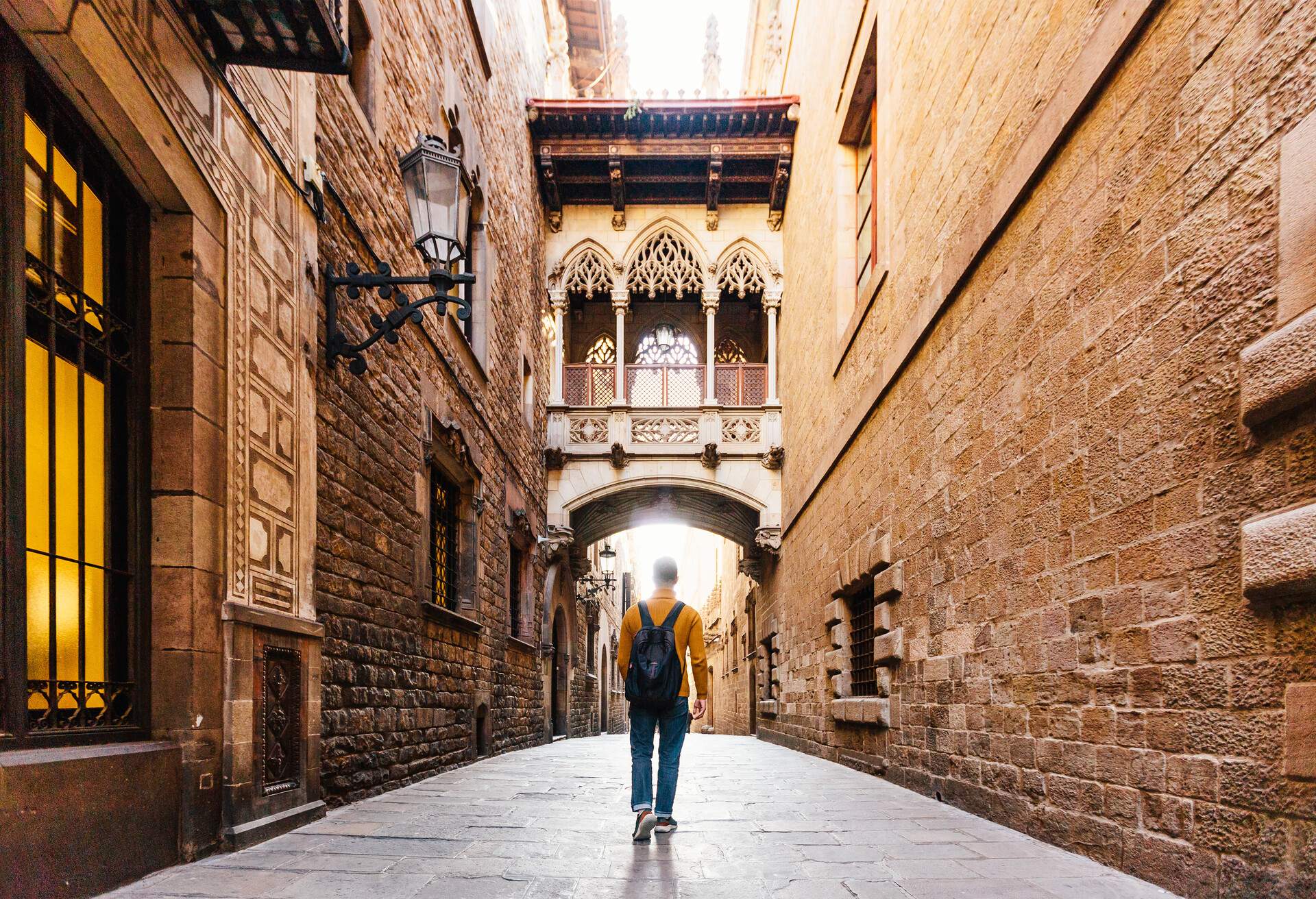 A man in an orange sweater and a black bag walks down a narrow alley with a small bridge in the middle.