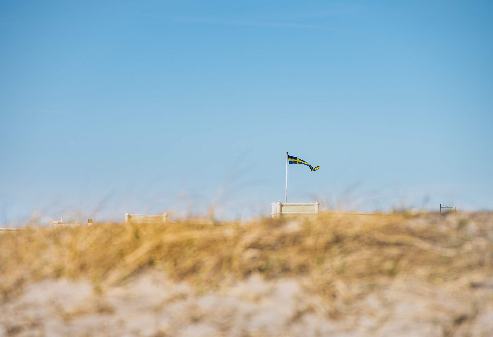 Swedish flag fluttering on a pole, captured through the sandy dunes of Ribersborgsstranden (Ribersborg Strand or Beach). Scandinavian Swedish banderole waving on Ribersborgs Kallbadhus - Malmo, Sweden