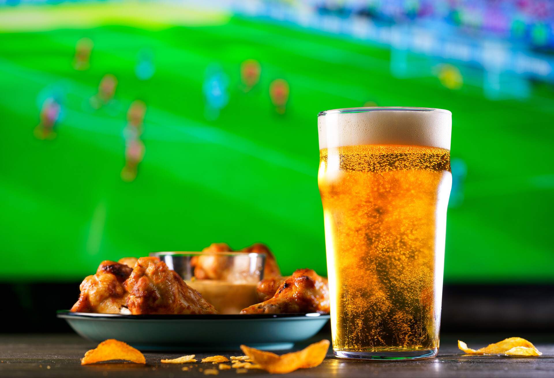 A glass of beer and hot chicken wings on a dark wooden table with crushed potato chips. Football on a background, high resolution; Shutterstock ID 1125372422