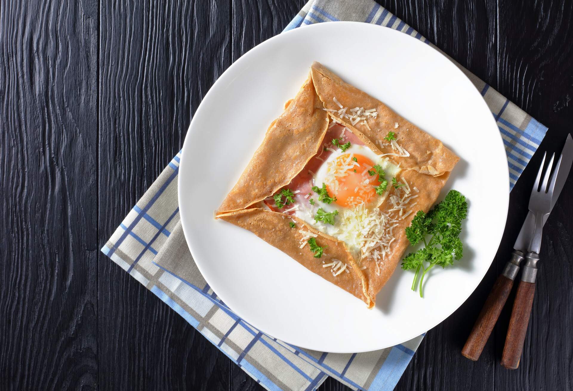 Breton crepe, Savory Buckwheat Galettes Bretonnes with fried egg, cheese, ham served on a white plate on a wooden table, french cuisine, view from above, flatlay, free space