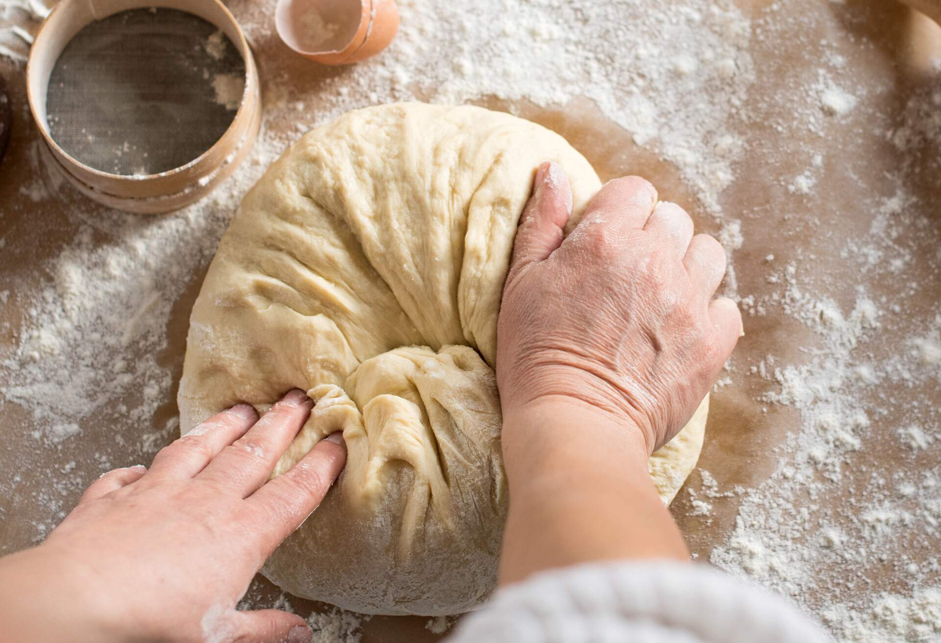 A pair of hands expertly kneading dough, with scattered flour on the work surface.