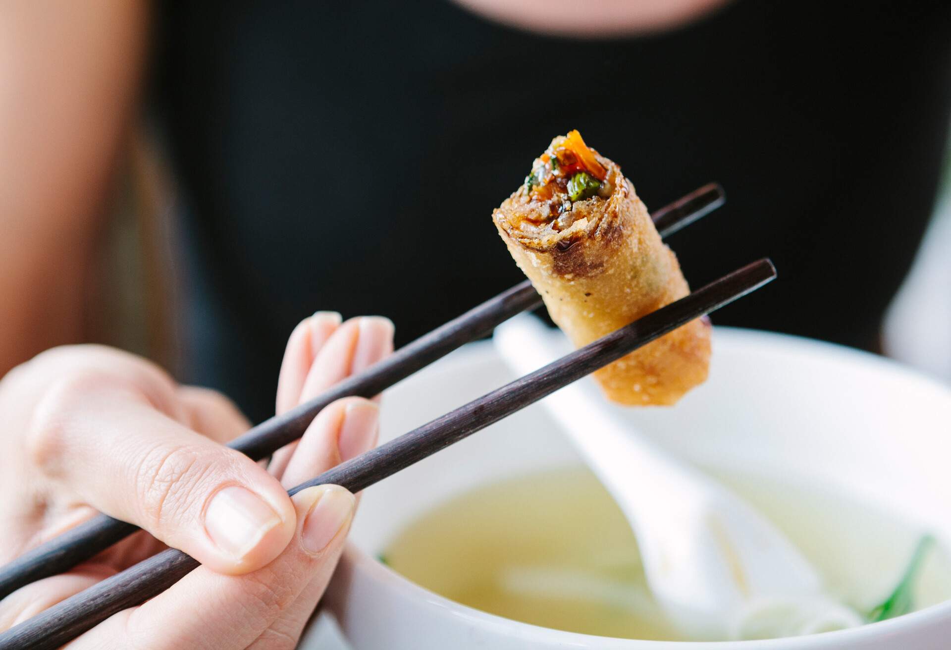 Chinese brunch - dim sum. Hand holding spring rolls with chopsticks. Wonton soup in the background.