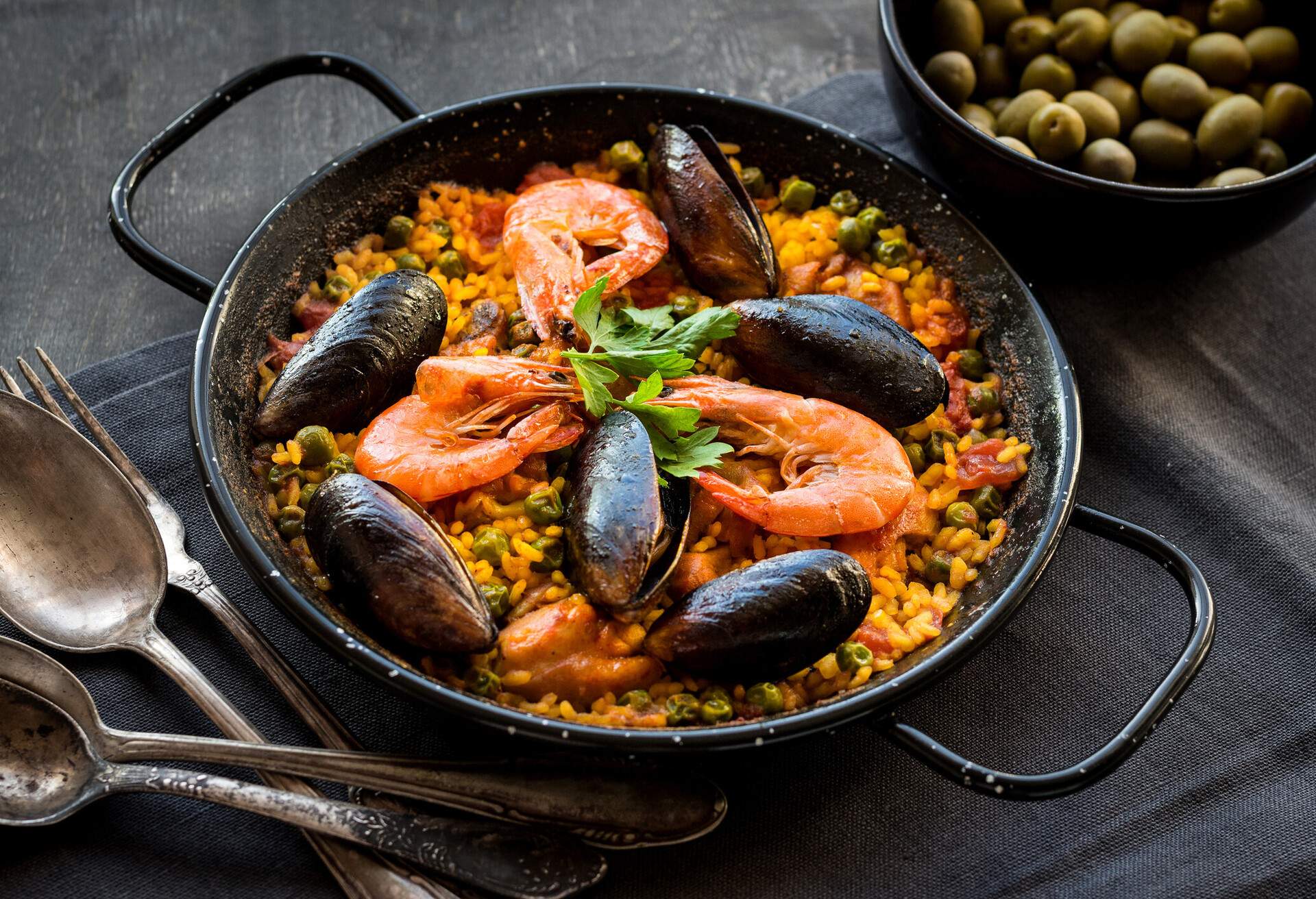 Paella in black pan with rice, shrimps, mussels, squid and meat, bowl with olives and vintage cutlery. Seafood paella, traditional spanish dish. Paella on rustic black wooden table. Selective focus