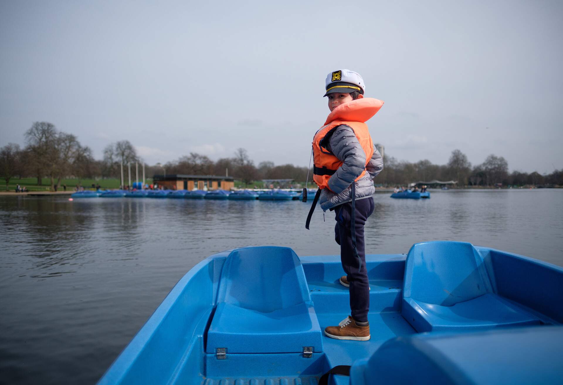 A young boy wearing a life jacket and sailors hat standing up at the prow of a pedalo boat on The Serpentine lake in Hyde Park, London.
