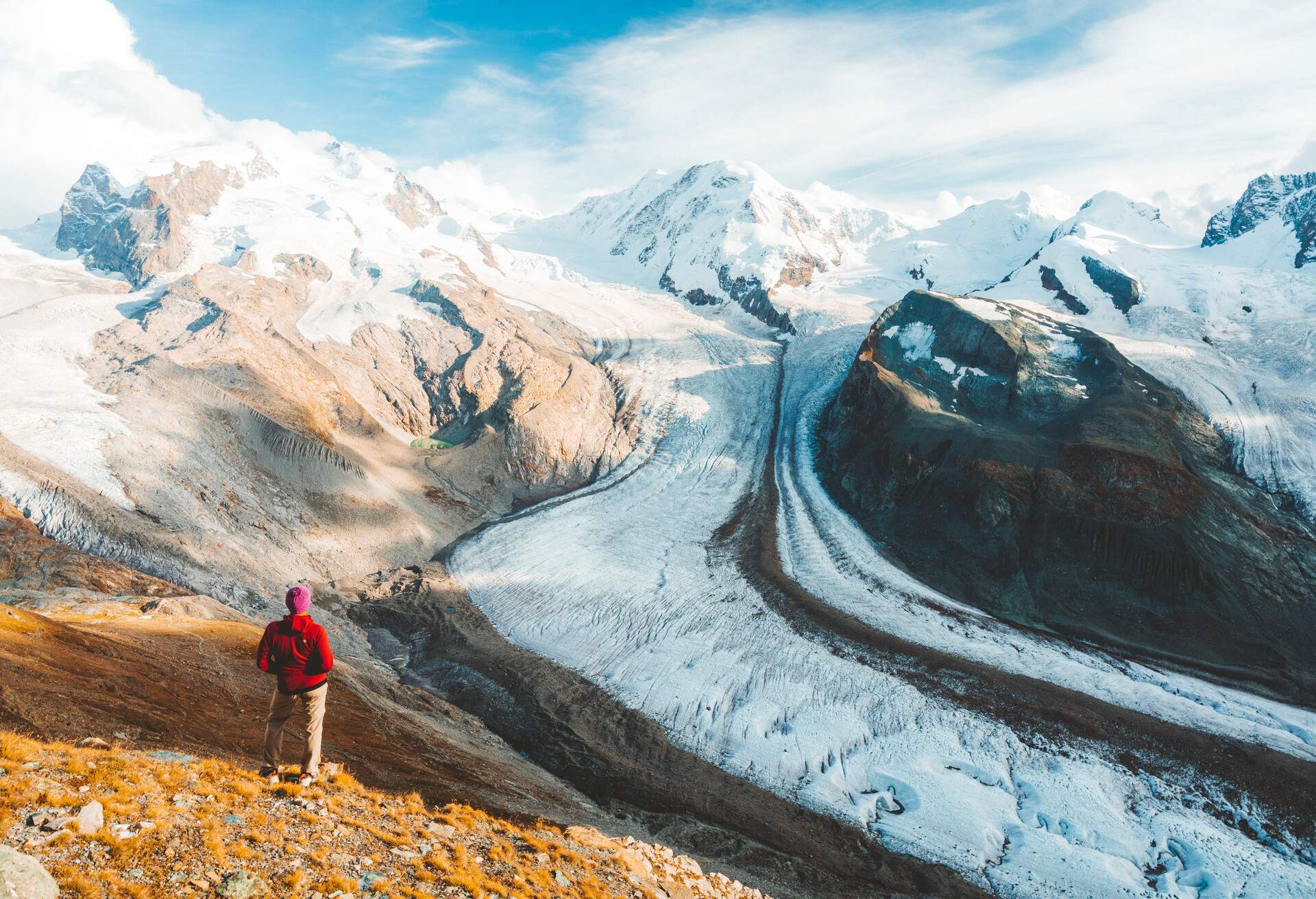 A hiker standing on a mountainside above a valley glacier looking towards distant snow-capped mountains.