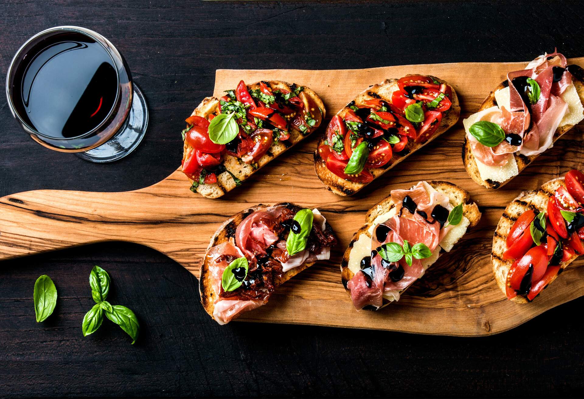 Brushetta set for wine. Variety of small sandwiches with prosciutto, tomatoes, parmesan cheese, fresh basil and balsamic creme served with glass of red wine on rustic wooden board over dark background, top view