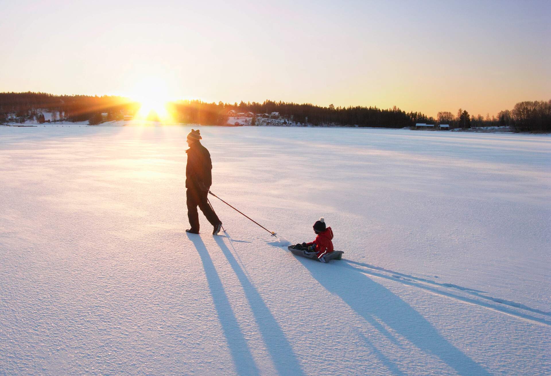The sun shines on a man pulling a boy on a sled in a snow field.