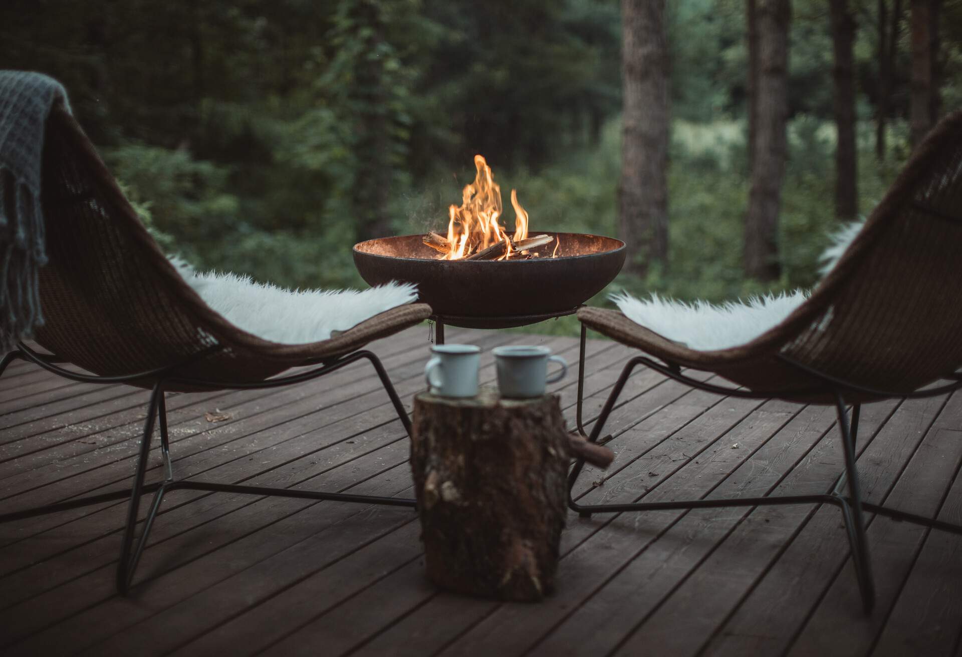A wooden porch with two cosy chairs around a campfire overlooking a lush forest.