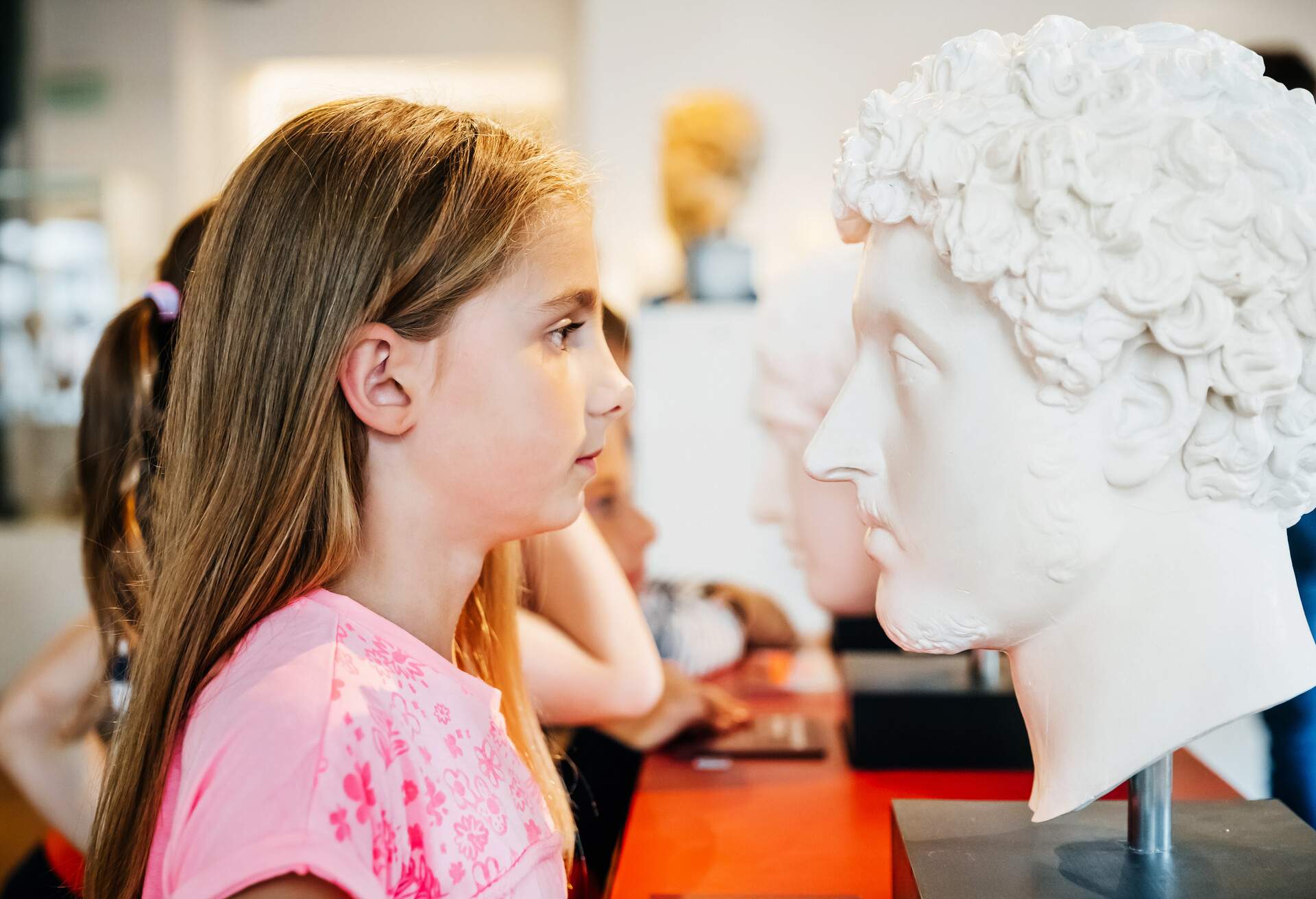 A young girl looking closely at a classical bust while in a museum on a field trip with her classmates.