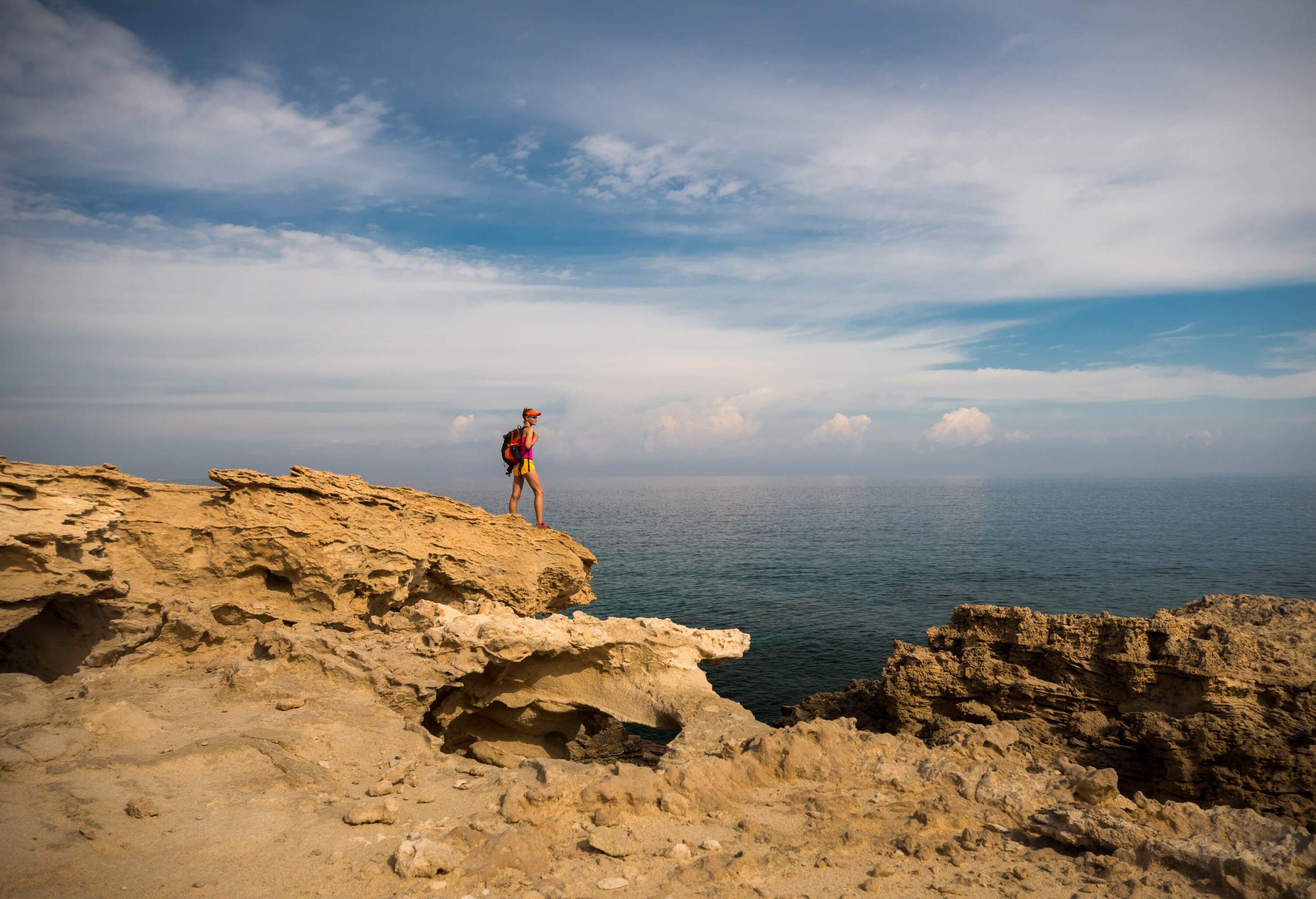 A male hiker with a backpack standing on a hanging cliff overlooking the vast ocean.