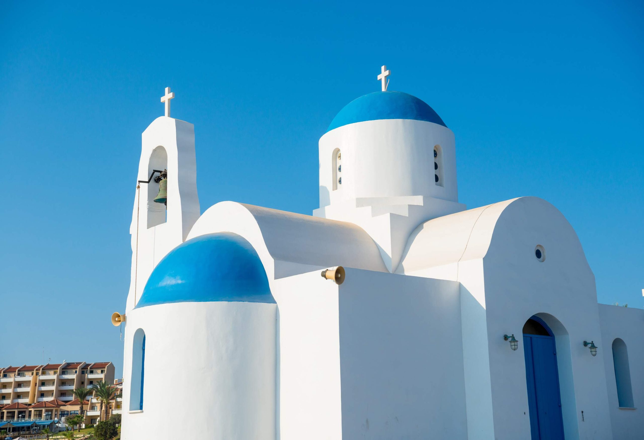 A white church with blue dome roofs and door, and a bell tower with cross finials.