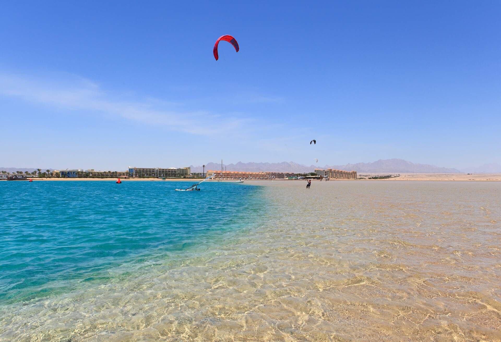 Two tourists kitesurfing on the shallow crystal clear waters of the bay with a row of buildings in the background.