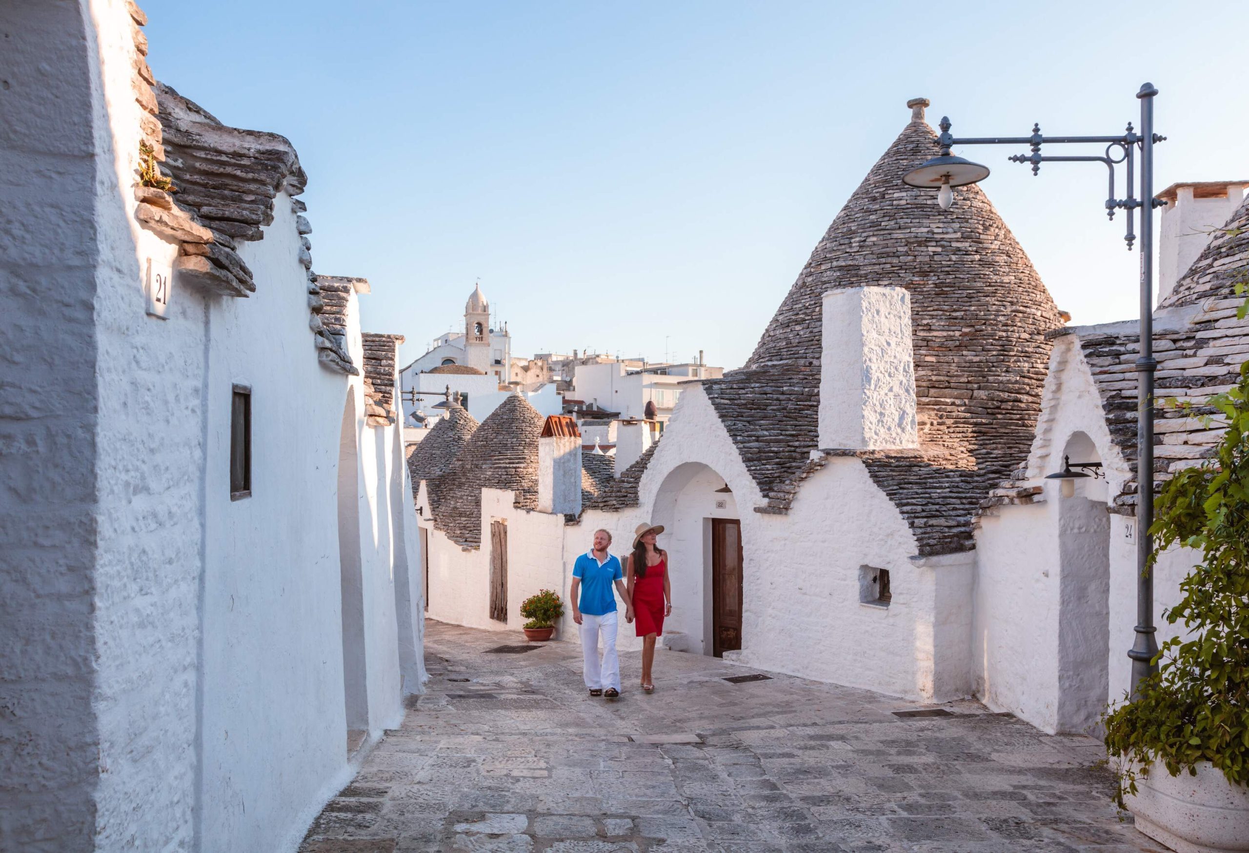 A couple holding hands while walking on a cobbled alleyway lined with trulli houses on both sides.