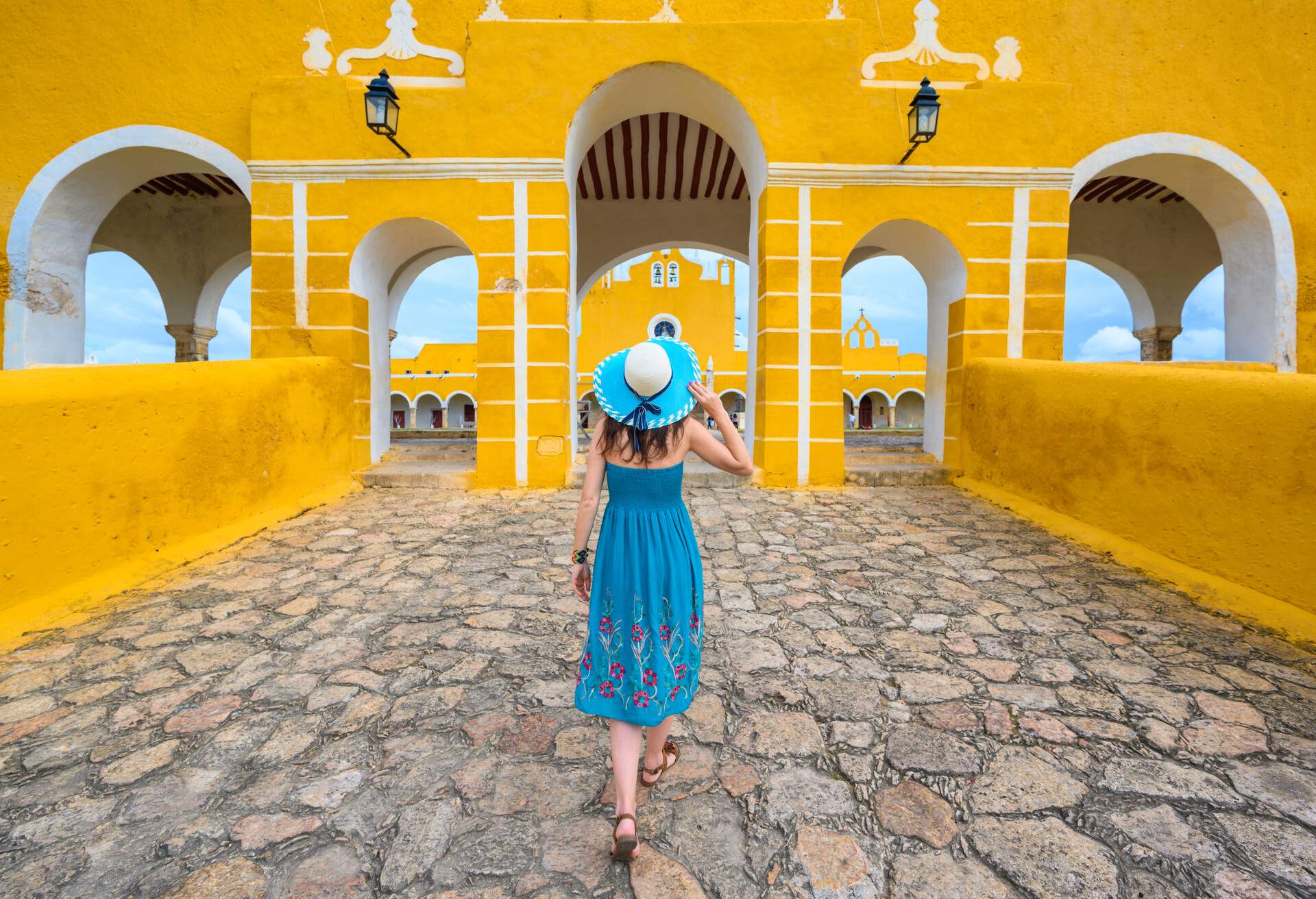 A woman in a blue dress and wide-brimmed hat walks towards a yellow building with multiple arch entrances.