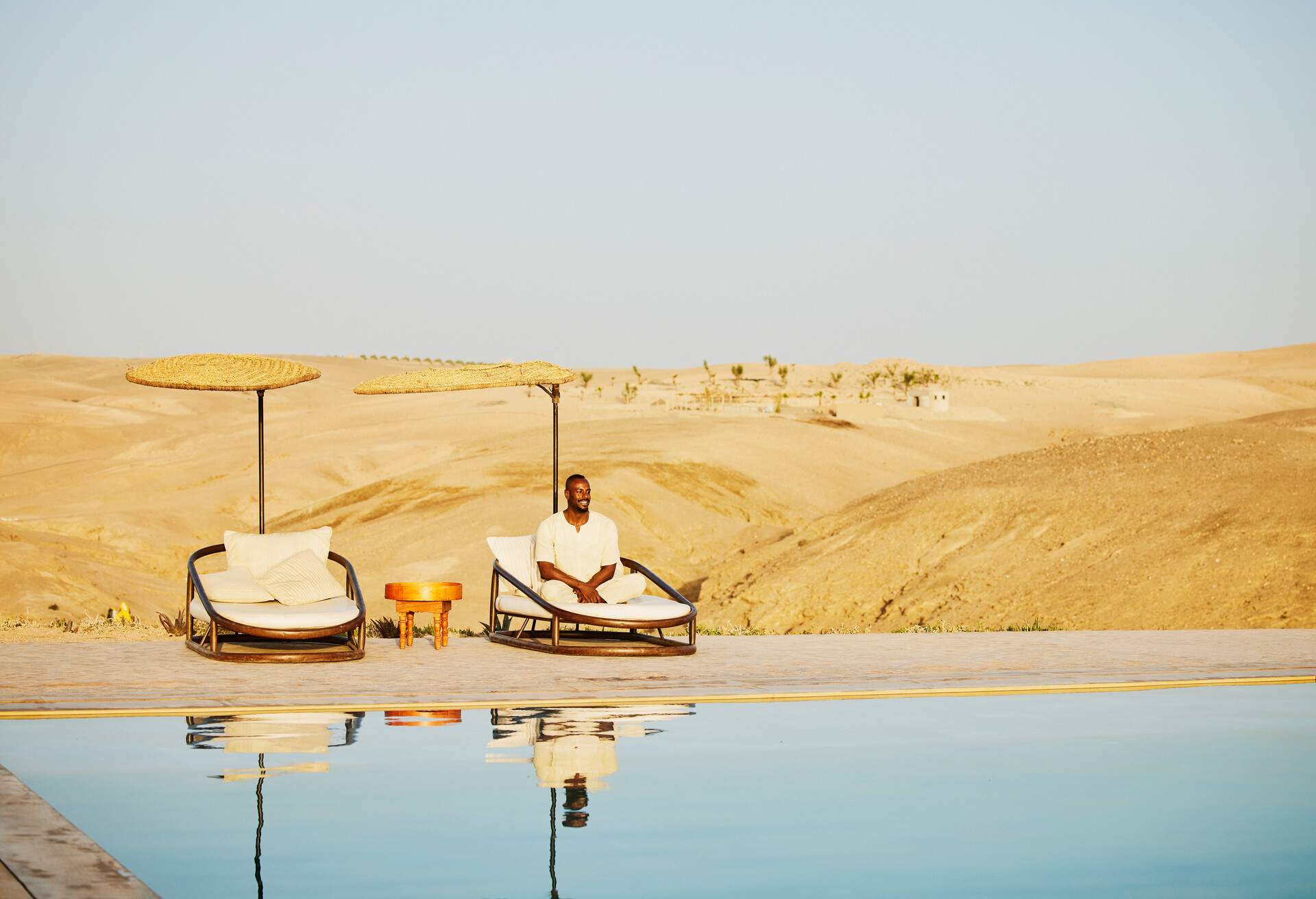 Wide shot of smiling man relaxing in lounge chair by pool and watching sunrise at Moroccan desert camp while on vacation