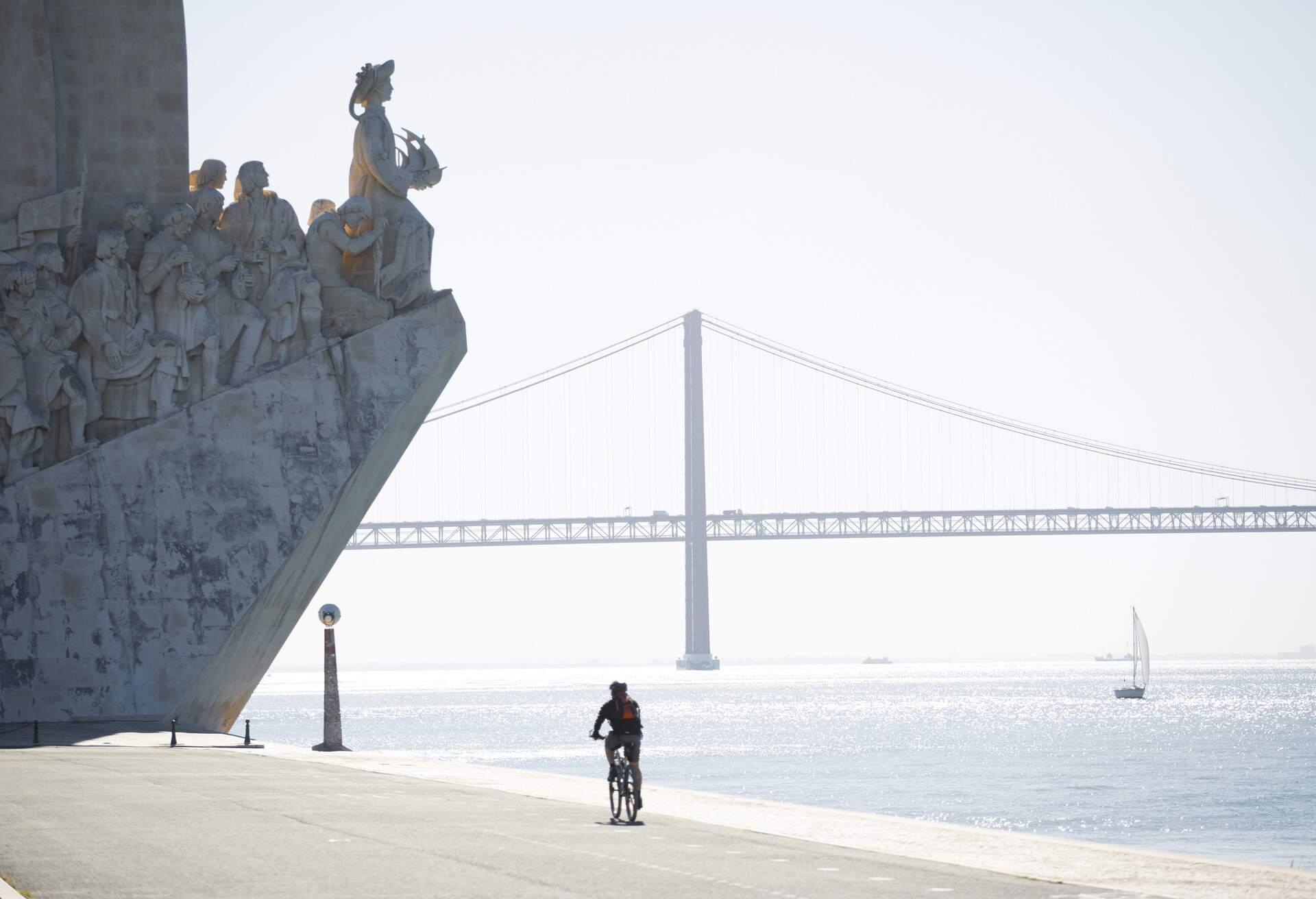 A cyclist approaches a riverside monument with statues of people going up a slope against a backdrop of a suspension bridge.