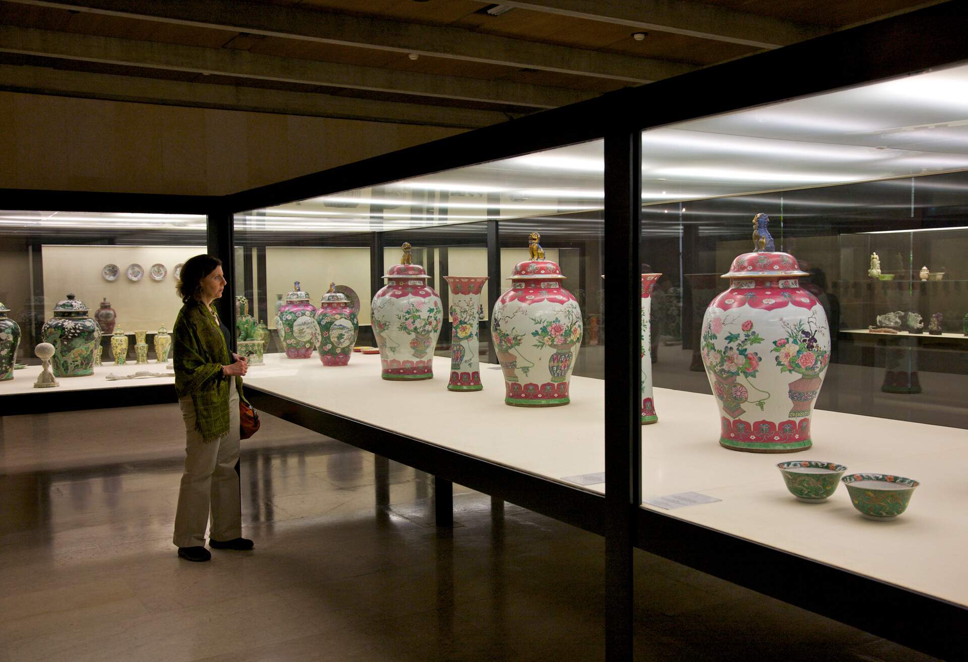 A mature woman looking at antique ceramic vases enclosed in a glass display case.
