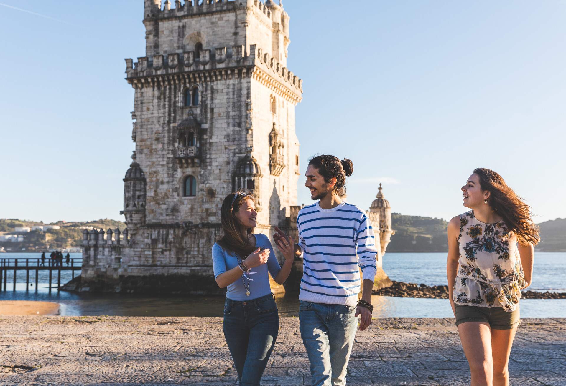 Three young people walking on a riverside promenade with the Belém Tower behind them.