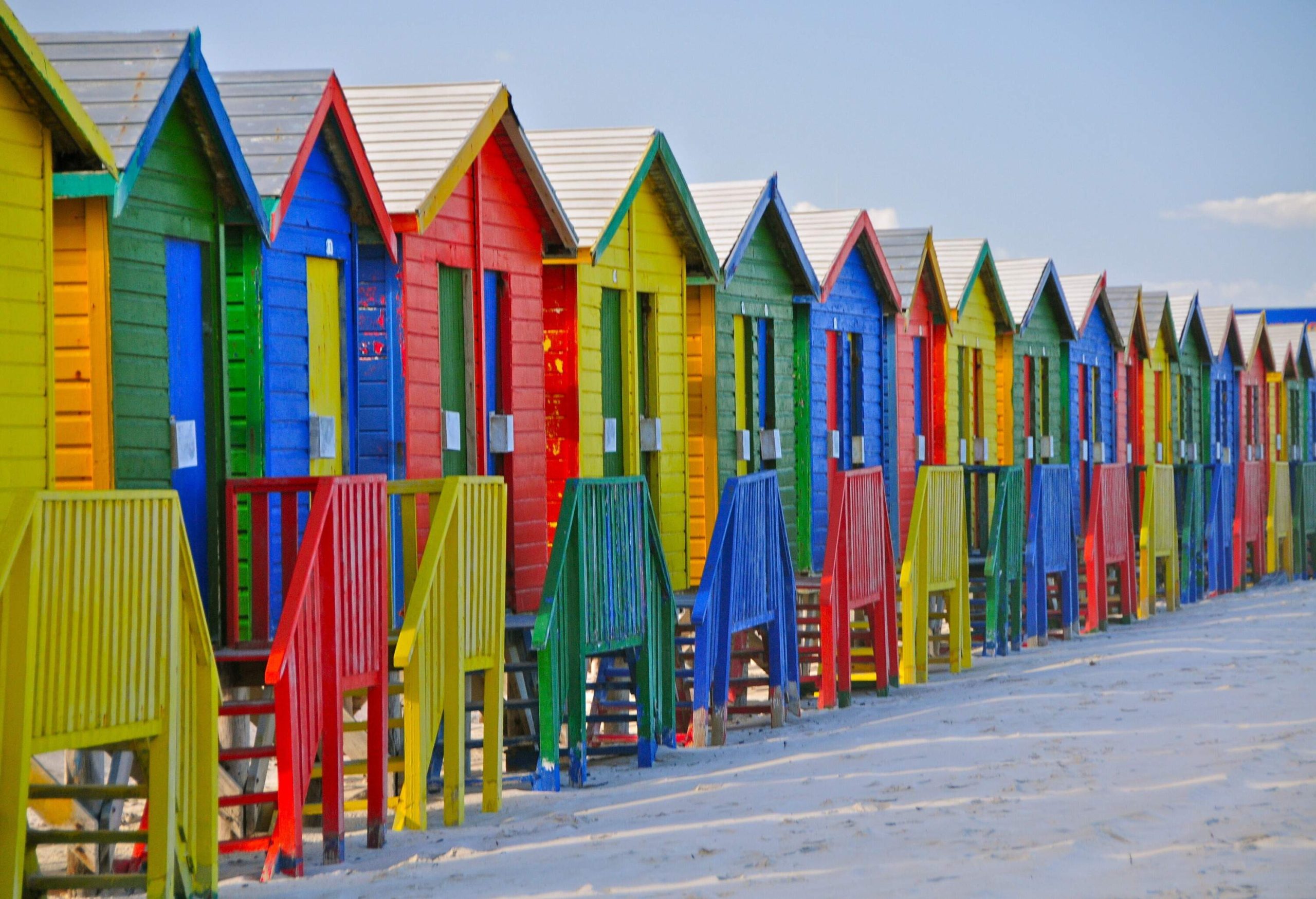 A series of rainbow-coloured beach huts lining up across the sand.