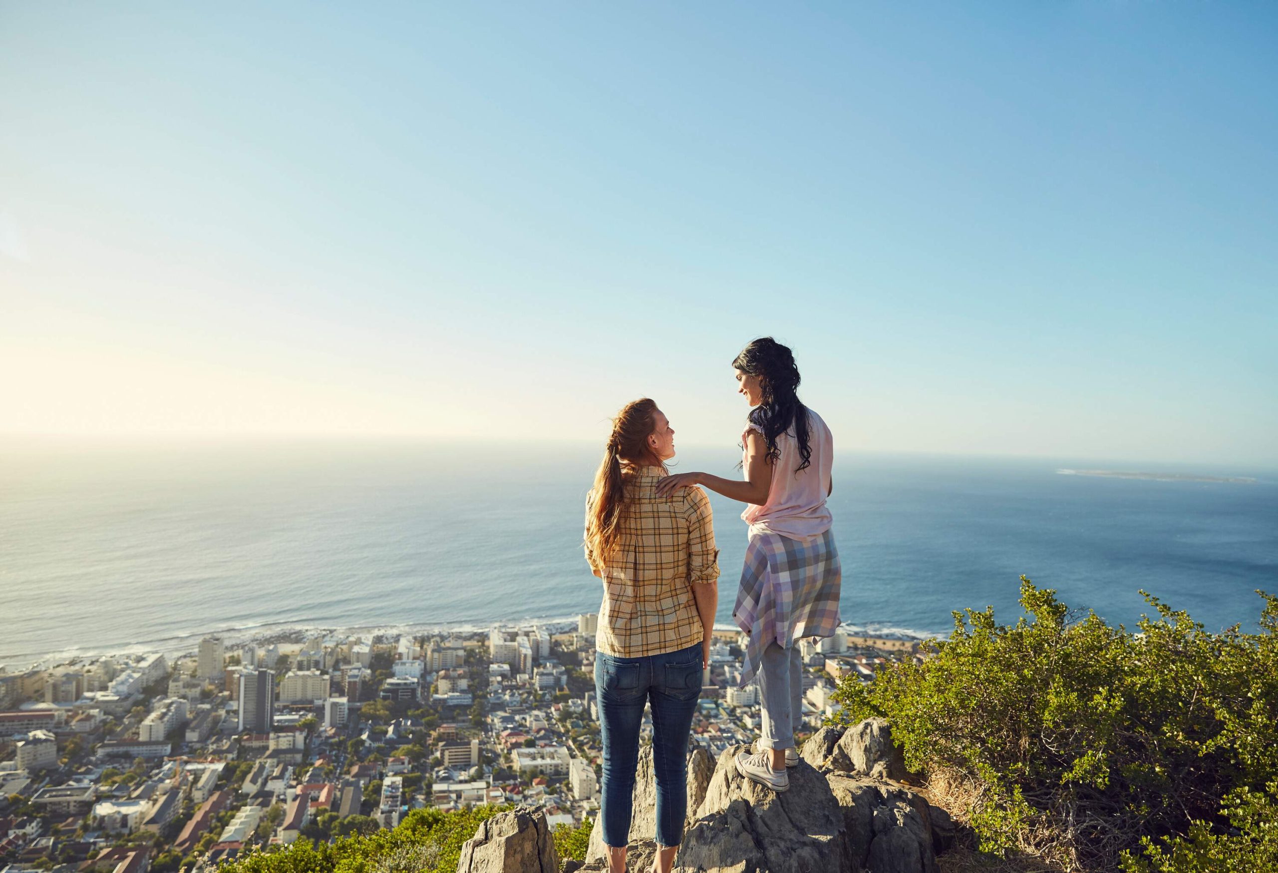 Two friends smile at each other, one placing her hand on the other's shoulder as they stand on a mountain peak with views of a seaside city.