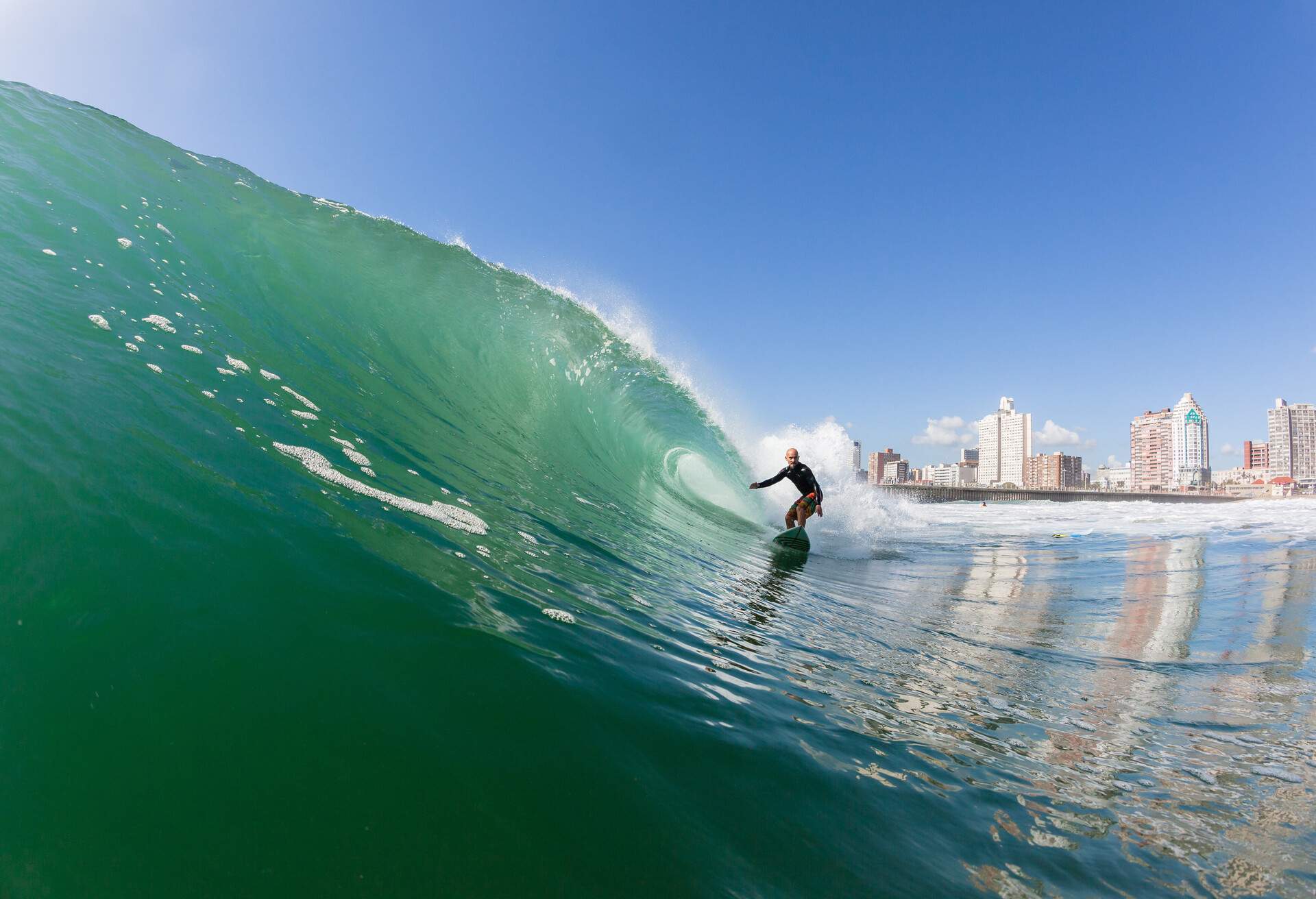 A surfer skillfully rides an ocean wave against the backdrop of modern tall buildings on the coast.