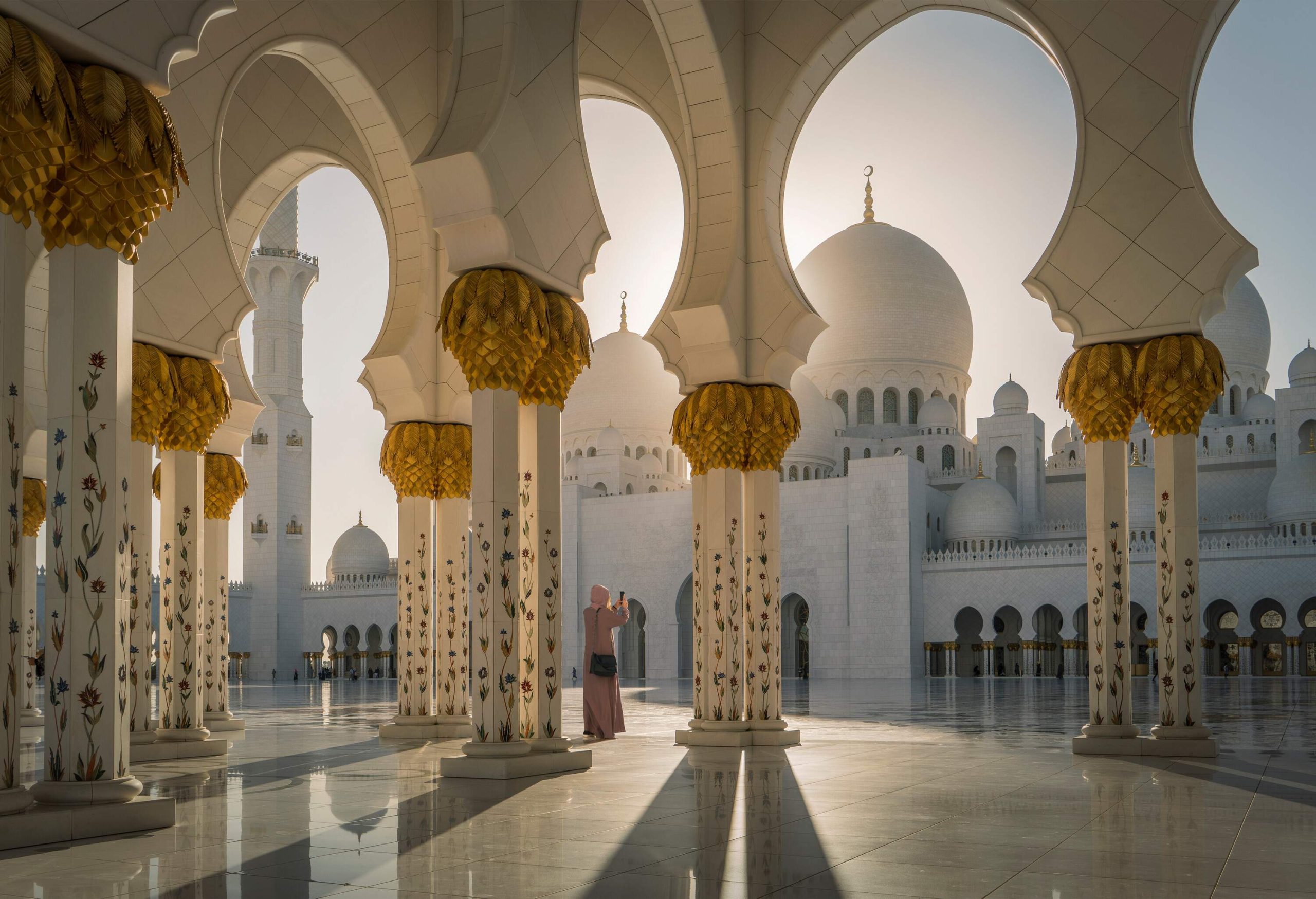 A woman in a robe standing inside the Sheikh Zayed Grand Mosque, taking a picture of the courtyard.