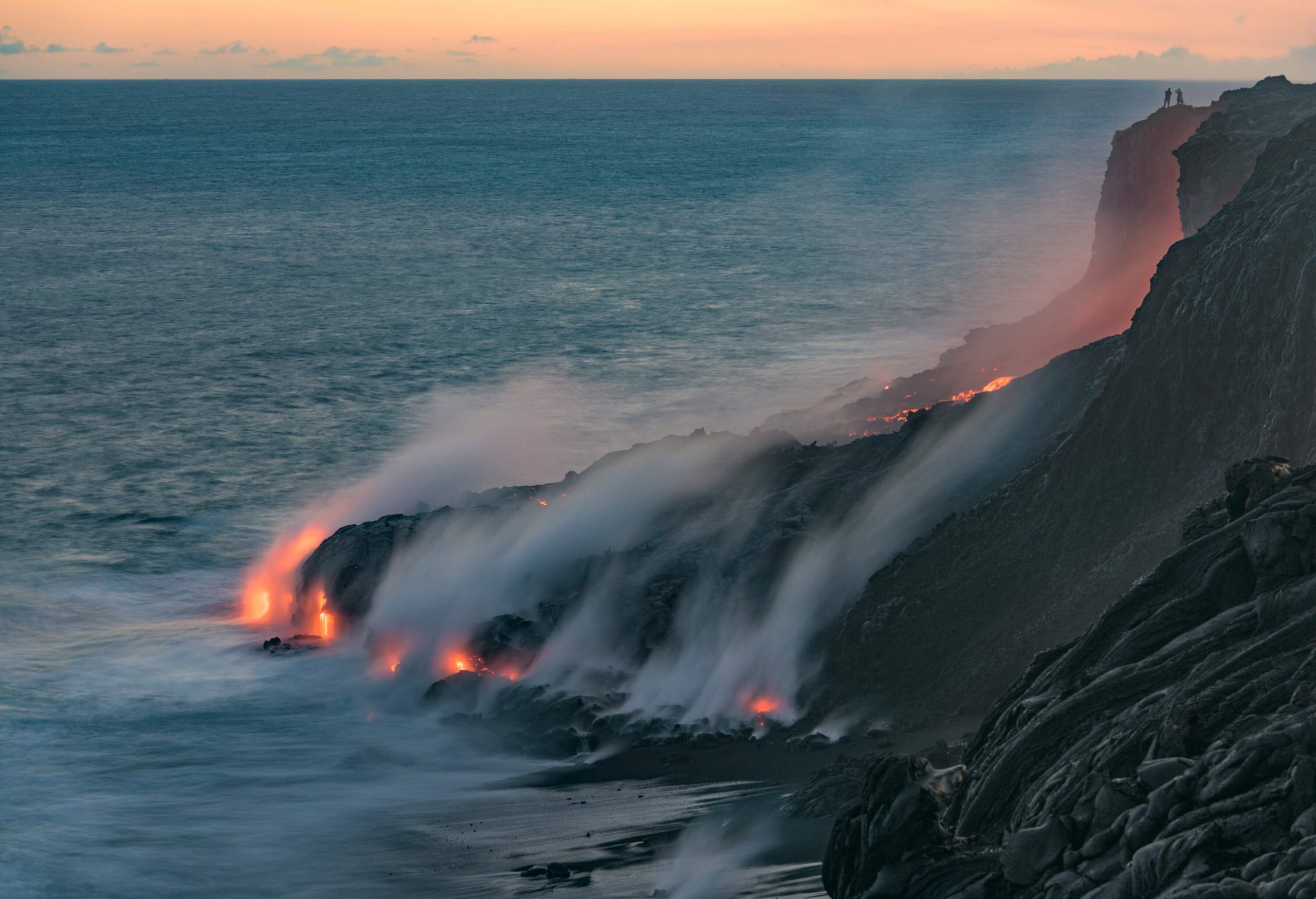 Molten lava flowing to the edge of the sea, emitting smoke as it touches the water, and a silhouette of two people standing on the peak.
