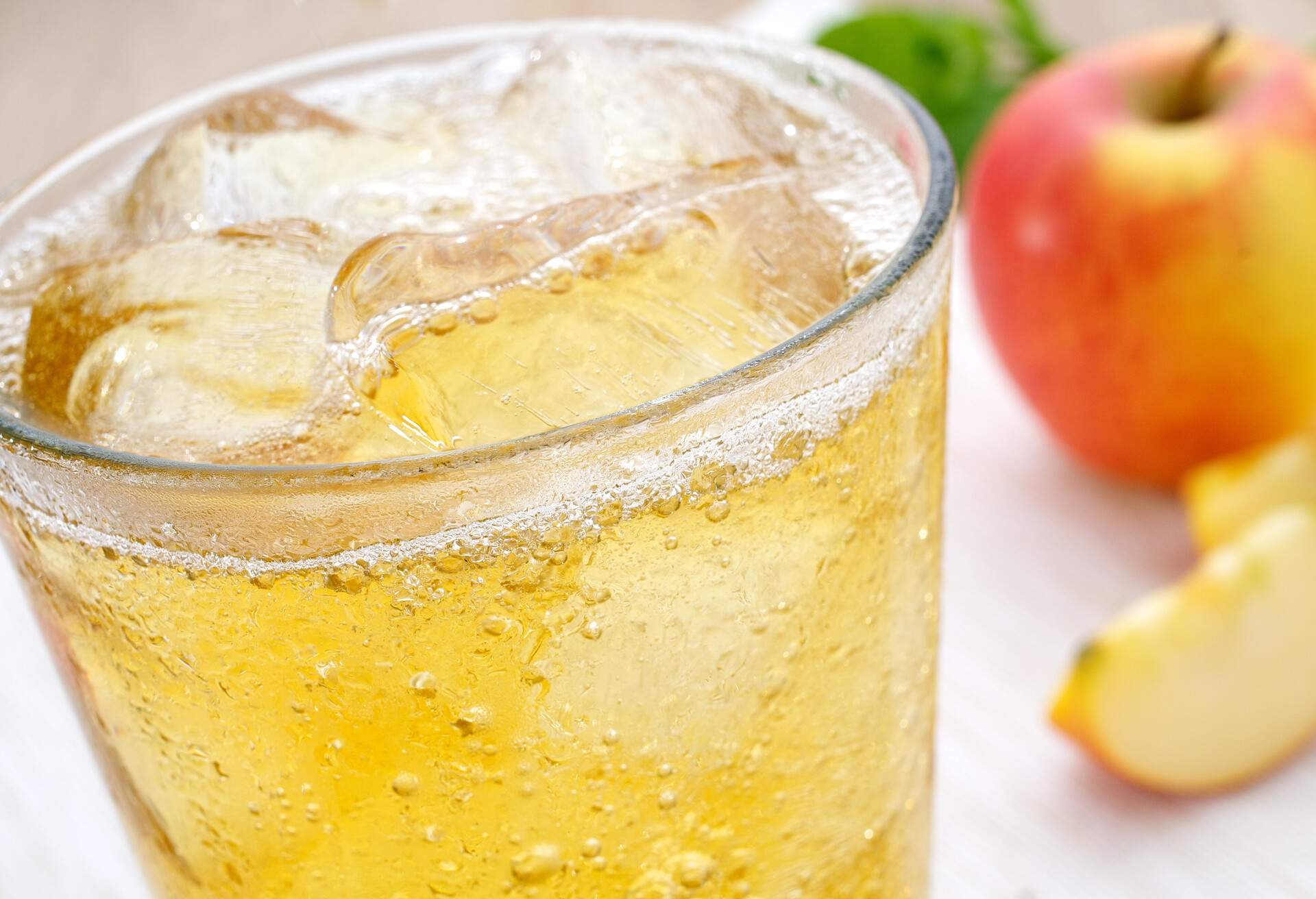 A glass of refreshing ice-cold apple juice spritzer apple spritzer with ice cubes. The cold glass has condensed water pots. In the background is an apple and sliced apple slices.