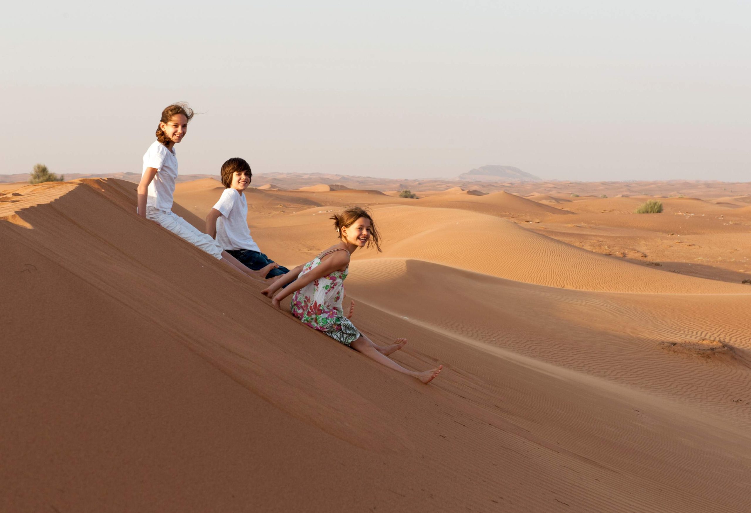 Three kids sliding down a mound of sand in the desert.