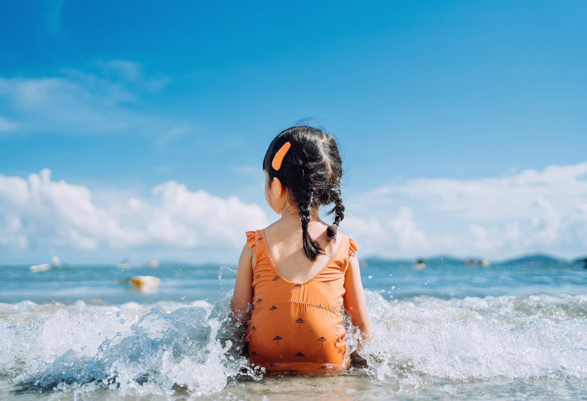 Rear view of lovely little Asian girl sitting by the seashore at the beach and being splashed by waves. Having fun at beach on a sunny Summer day