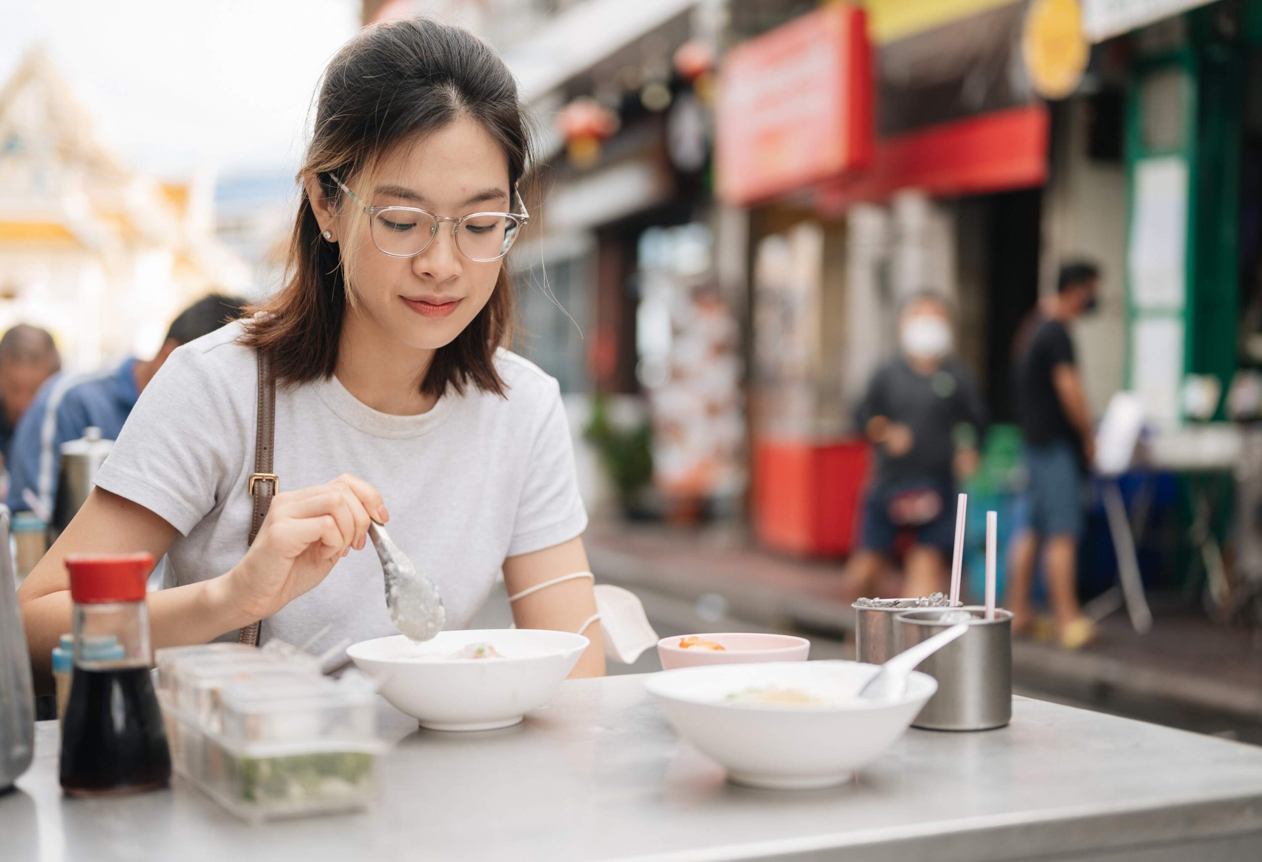 Woman in a white shirt and eyeglasses sits on a table mixing at a bowl of porridge.