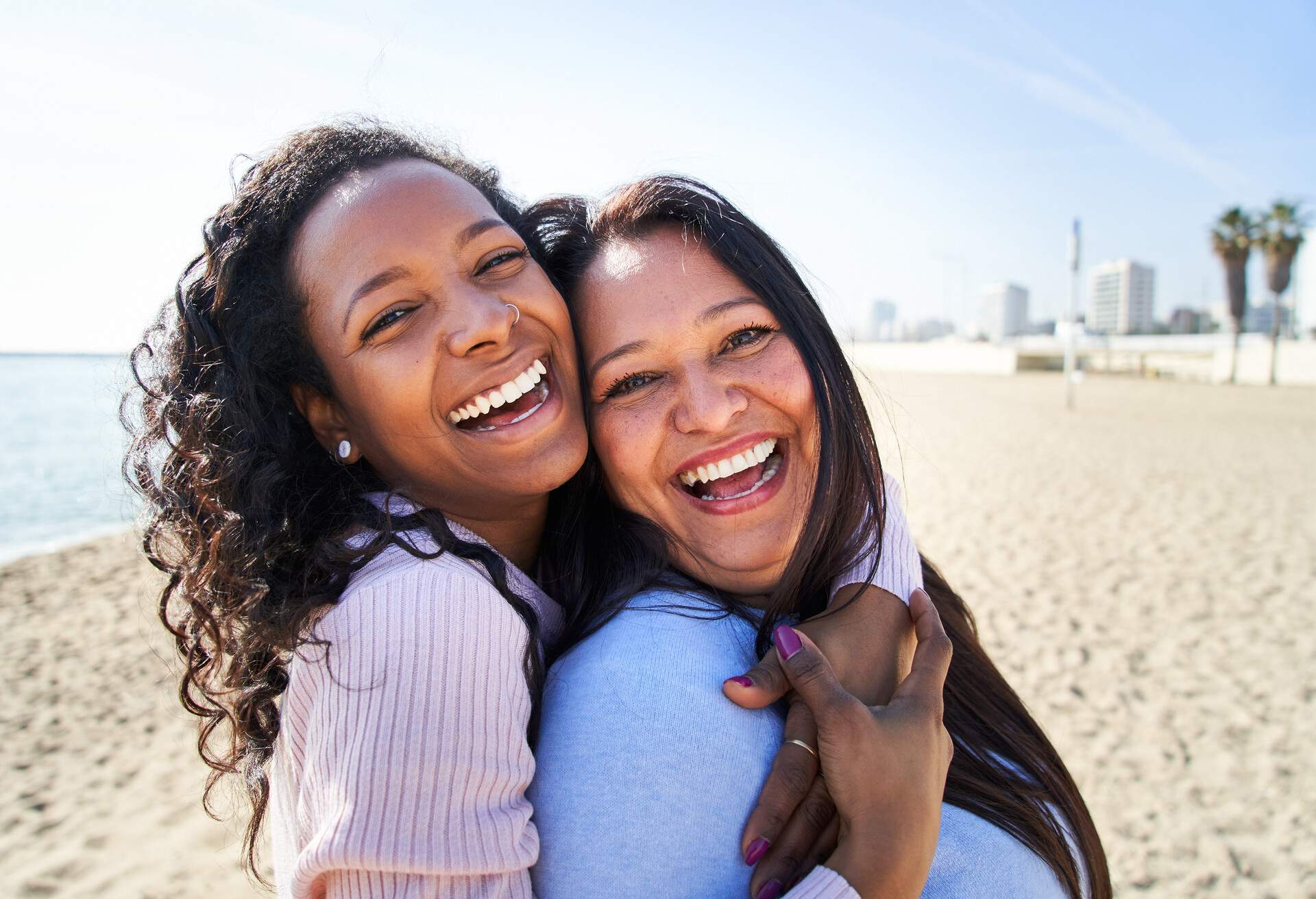 Close-up photography of multiracial family, looking and smiling at camera, daughter hugging her mother, standing on the beach, woth big smile.