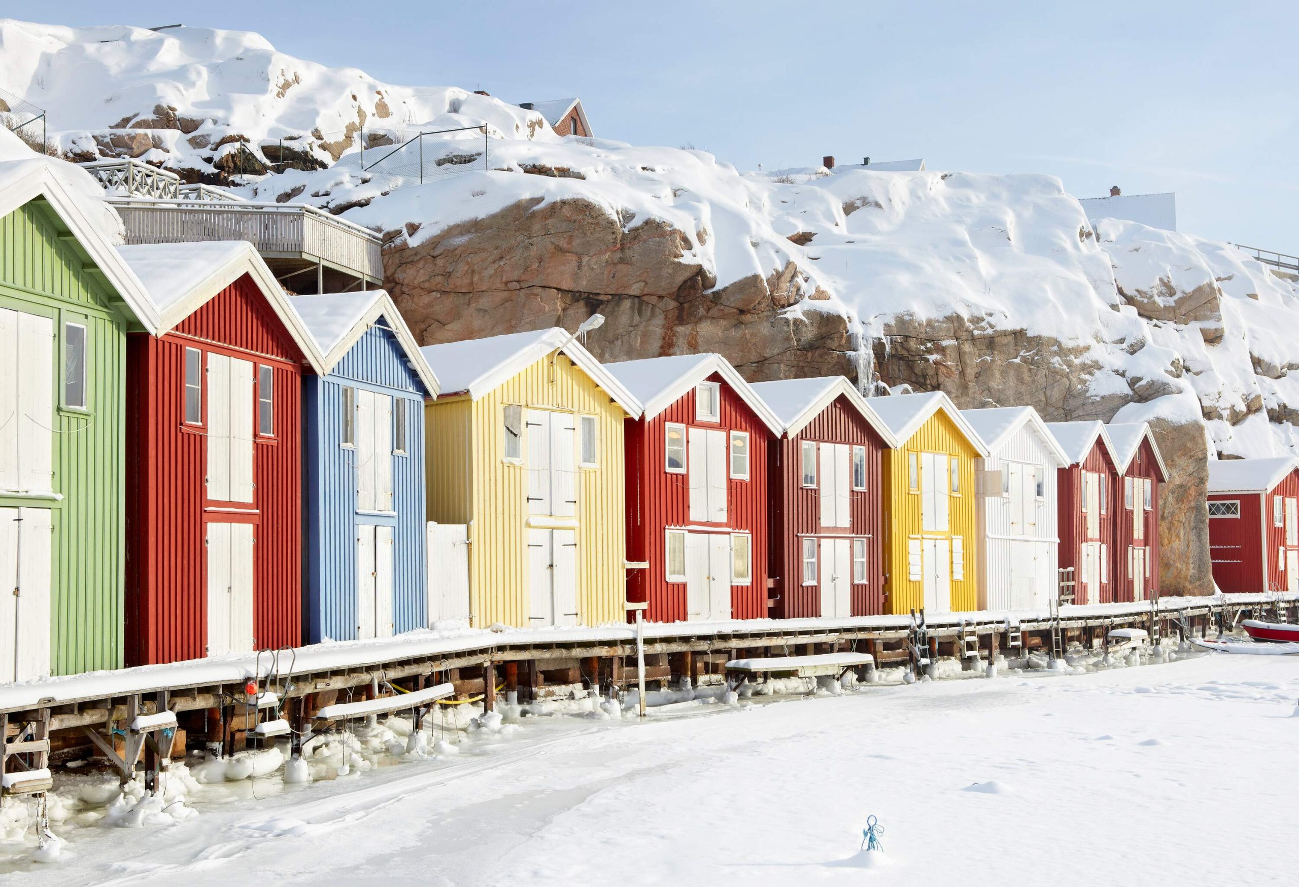 Row of colourful fishing cabins with hills covered with snow in the background.