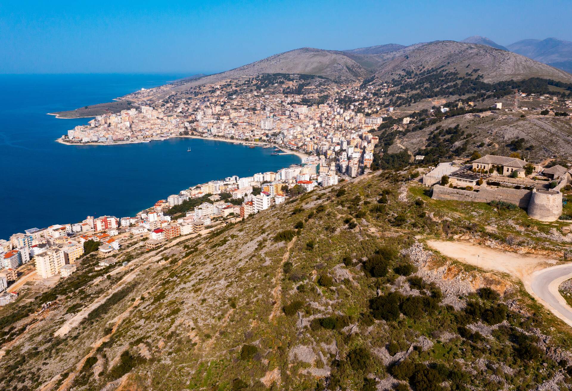 Scenic aerial view of Sarande cityscape on shores of gulf of Ionian Sea and ancient Lekuresi Castle on hill above city on sunny spring day, Albania