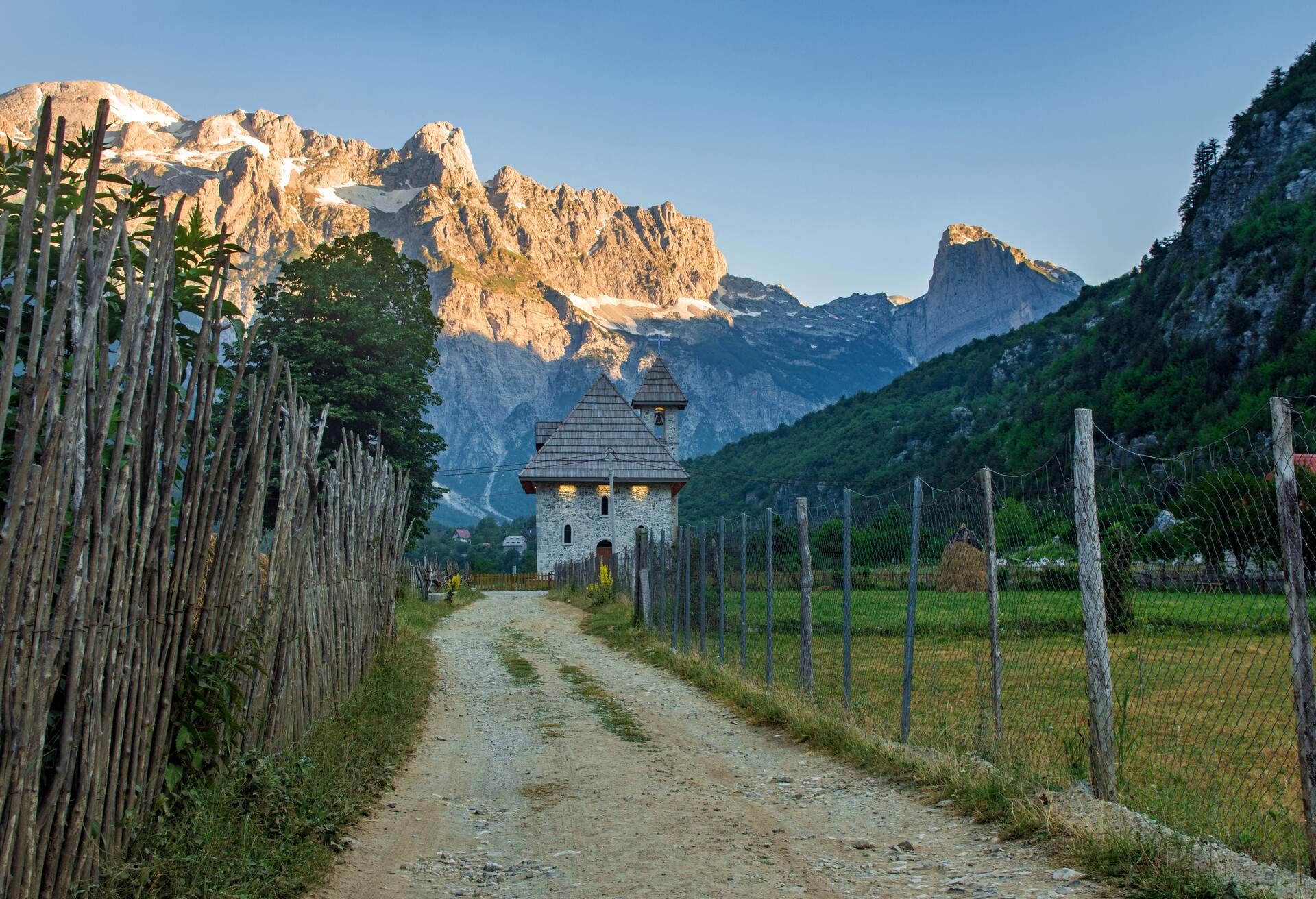 a path with wooden fence leads the catholic church in the Theth Valley, on background the mountains of Albanian Alps