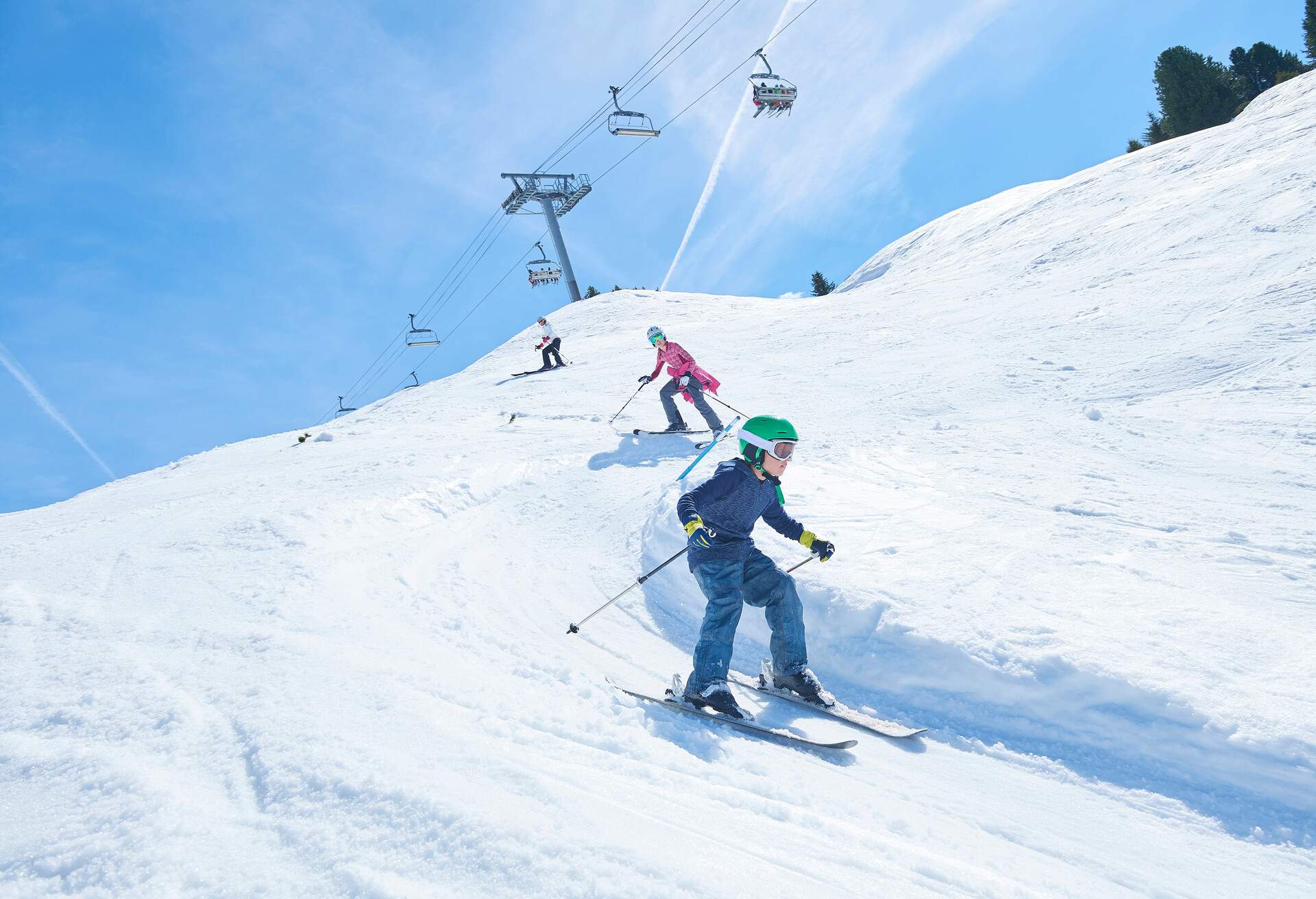 Young kids skiing down a slope with chairlifts passing overhead.