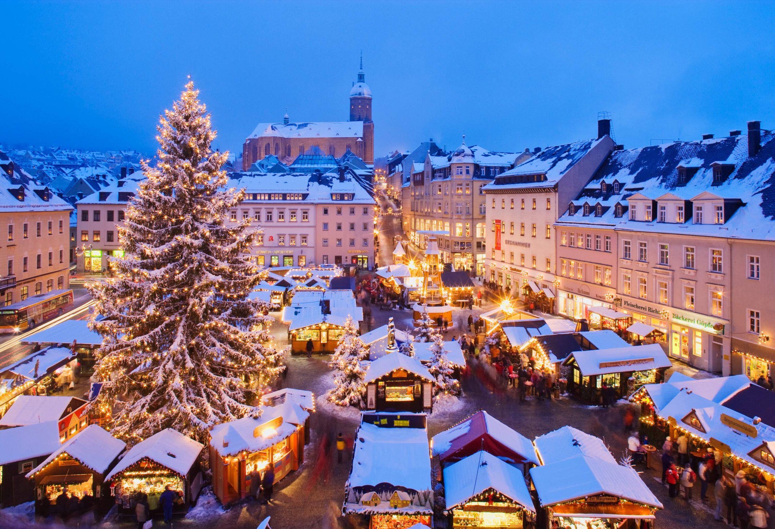 A busy Christmas market with a large Christmas tree in the middle surrounded by city structures at night.
