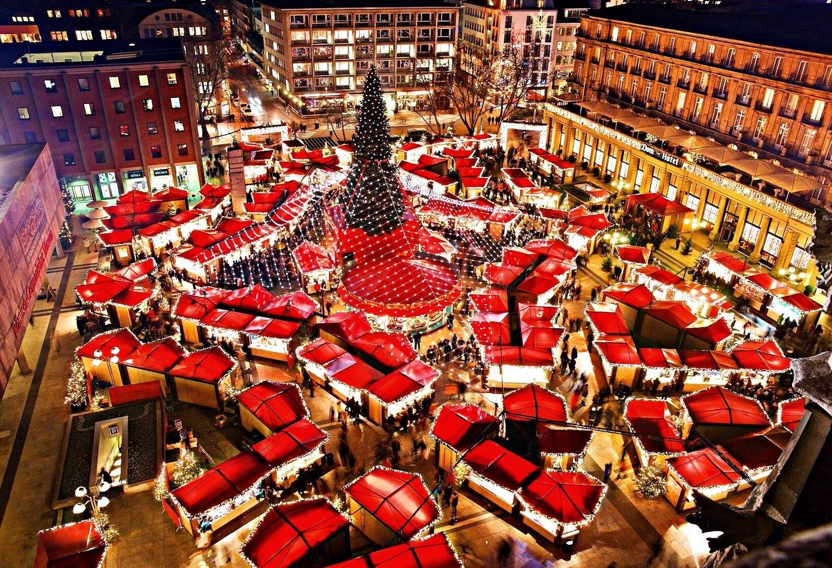 Aerial view of a market with a tall illuminated Christmas tree in the centre surrounded by stalls with red roofs.