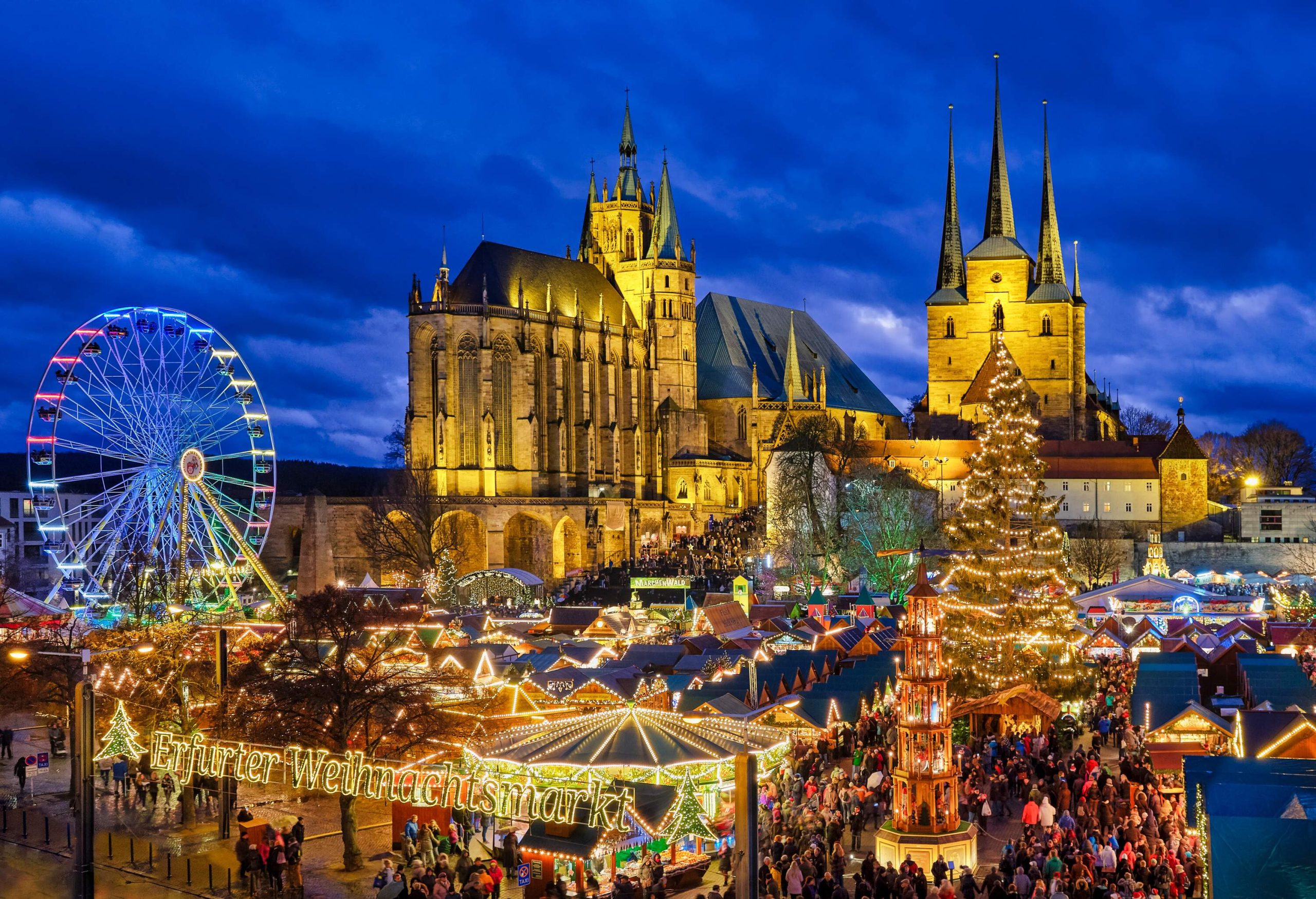 A lively Christmas market heavily decorated with holiday lights with marvellous Gothic structures in the background.