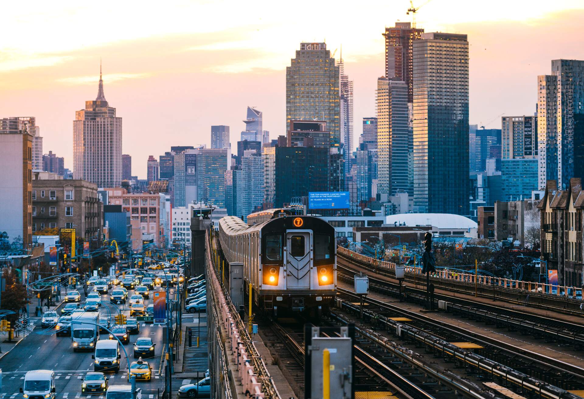 A train on an elevated railway running alongside a busy road backed by towering buildings.