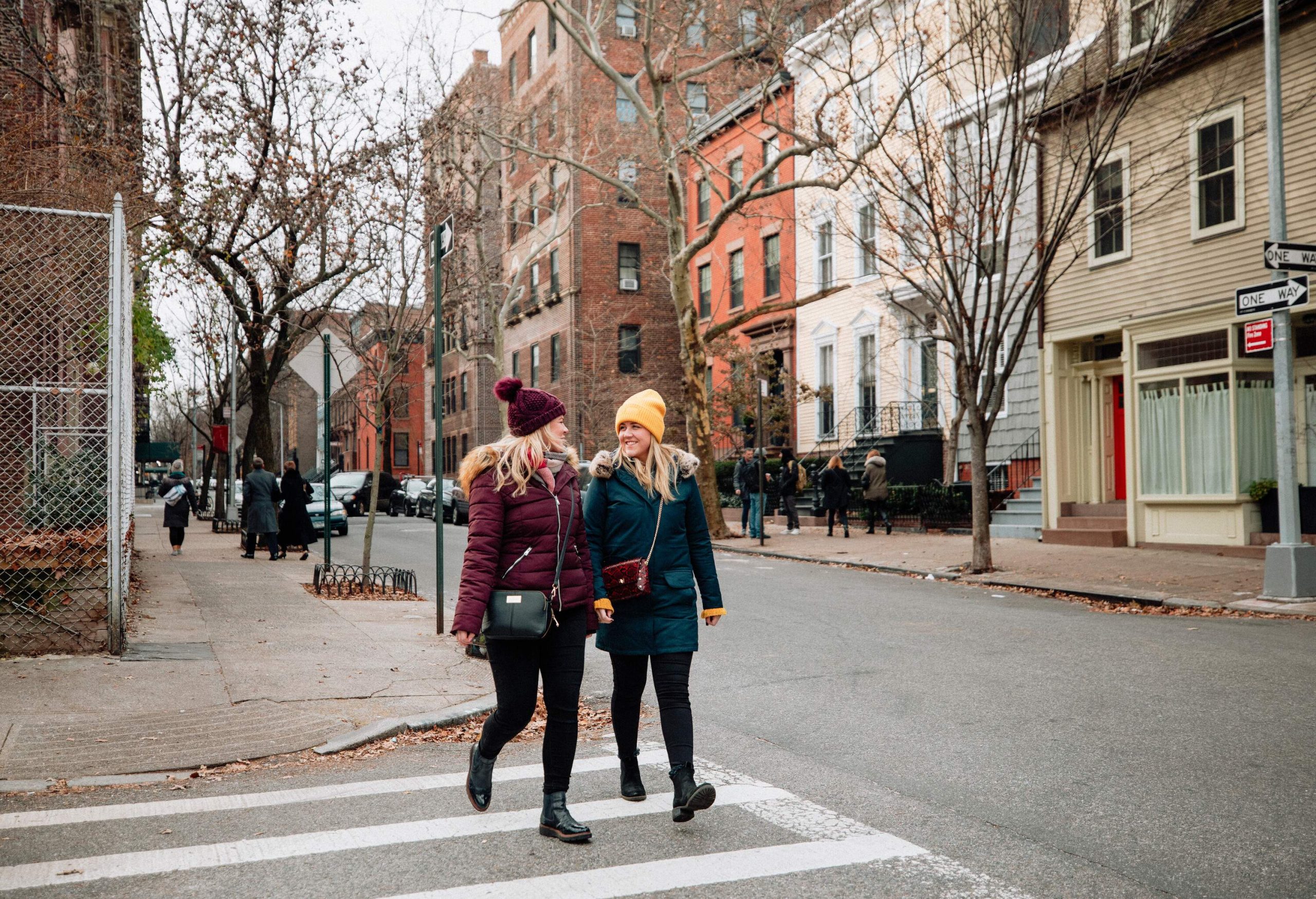 Two blonde women in winter clothes and knitted bonnets cross a pedestrian crossing in a neighbourhood with streets lined with bare trees.