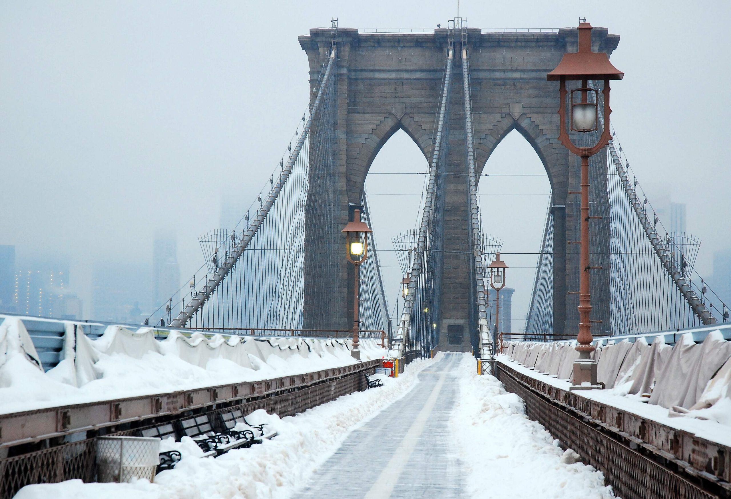 Snow covered walkway with lamp posts towards the hazy entrance of Brooklyn Bridge.